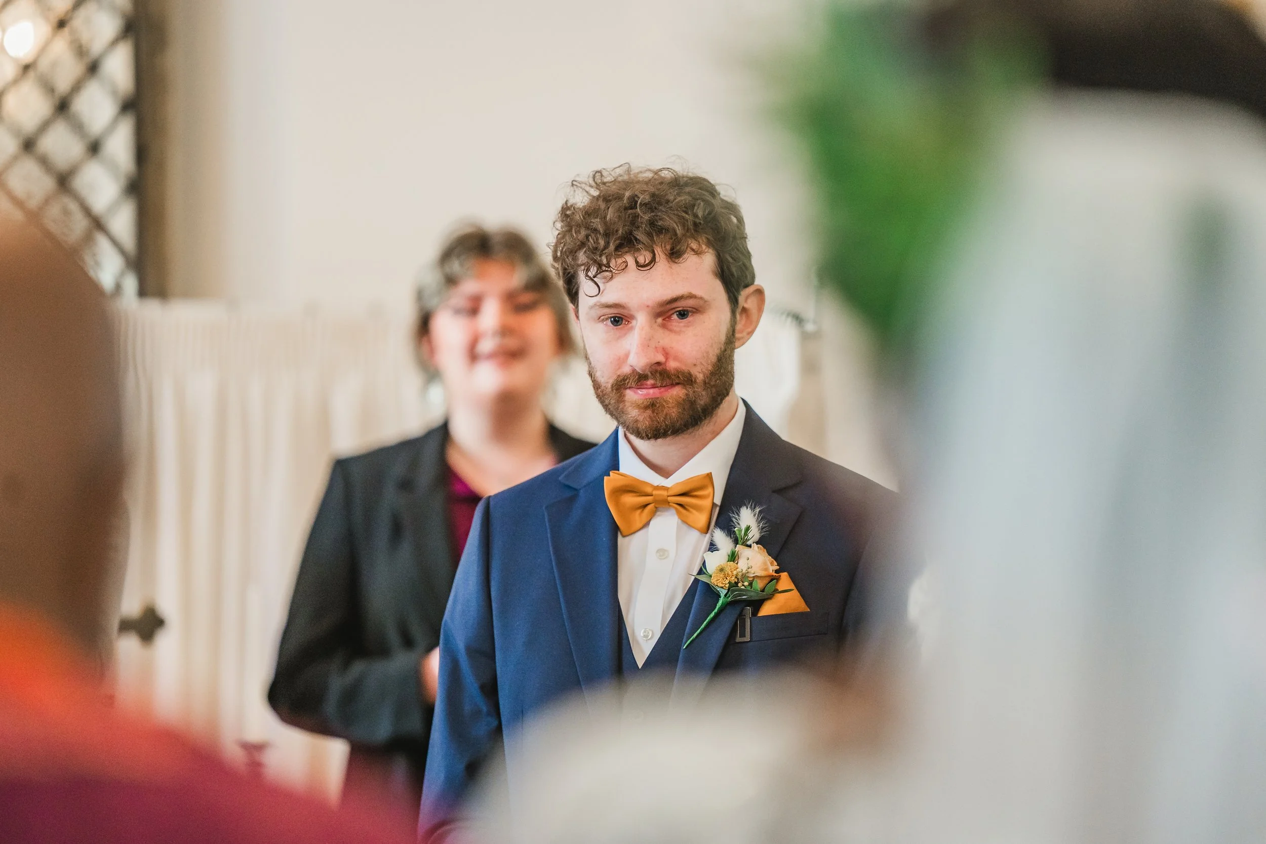 Young man with curly hair and beard wearing a navy suit with a yellow bow tie and boutonniere, standing in front of a mirror with blurred woman in background, possibly during a wedding or special event.