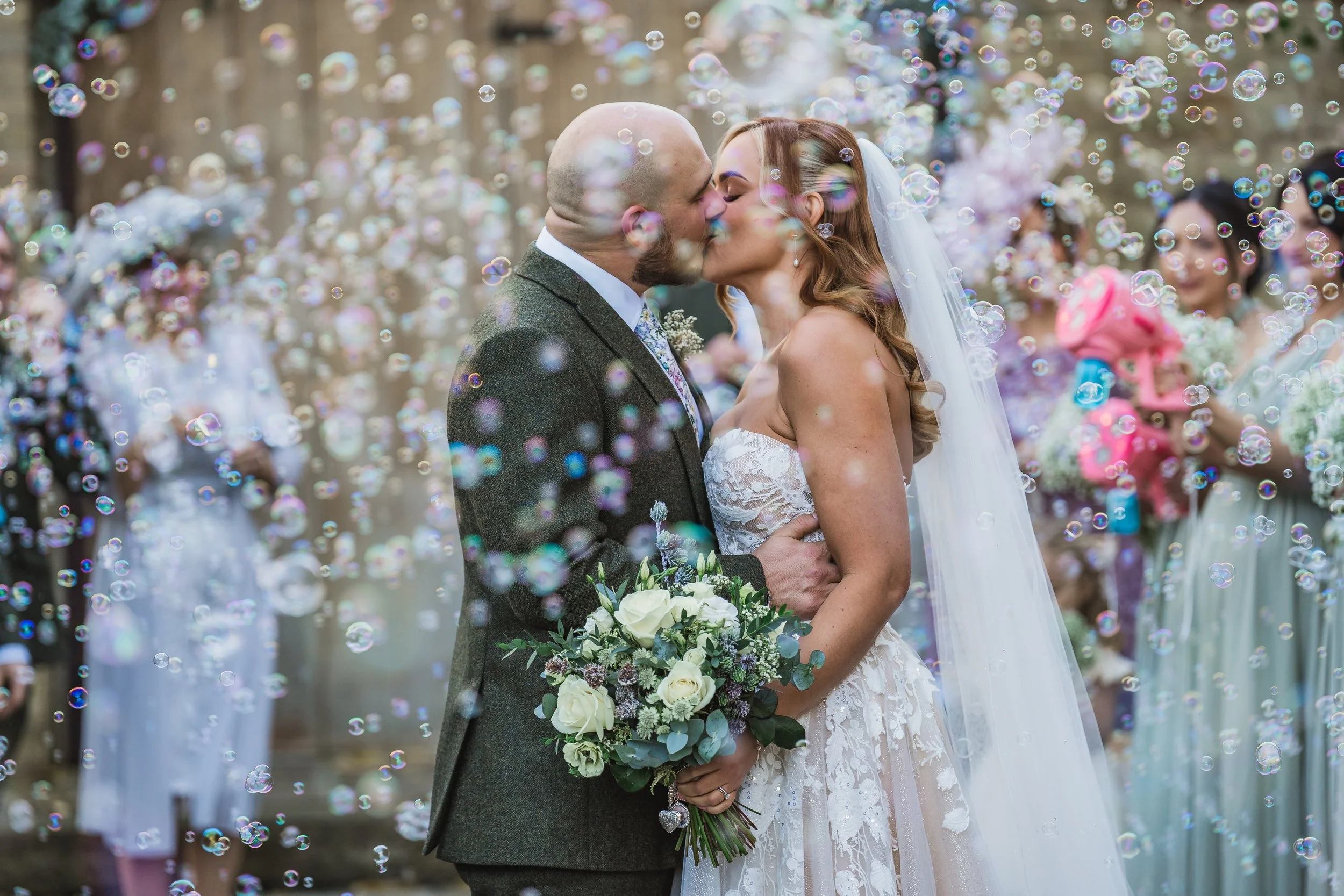 A newlywed couple shares a kiss at their wedding surrounded by friends, with bubbles floating around them.