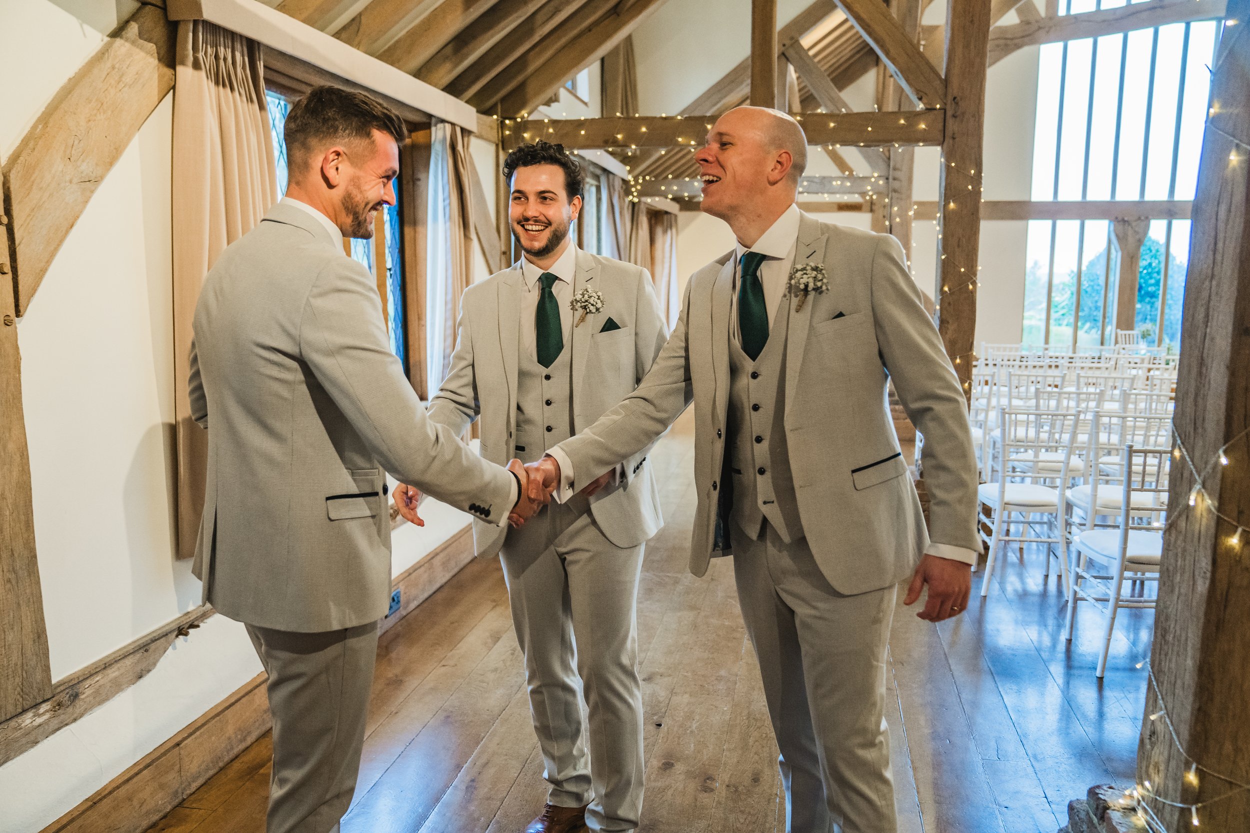 Three men in gray suits shaking hands and smiling at a wedding ceremony in a rustic venue with wooden beams, fairy lights, and rows of chairs.