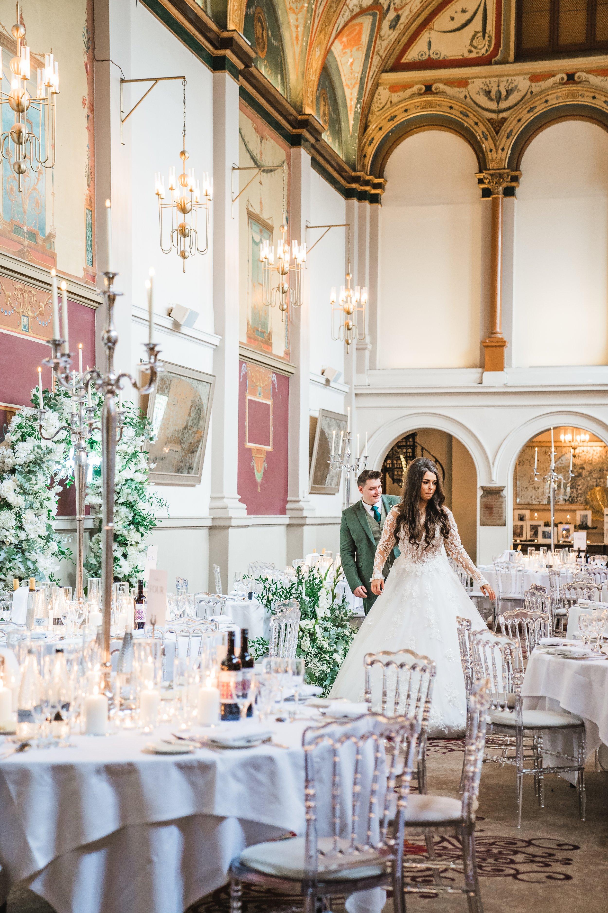 A bride in a white wedding gown with lace sleeves walking with a groom in a green suit inside an elegant decorated hall with high arched ceilings, chandeliers, floral arrangements, and set tables.