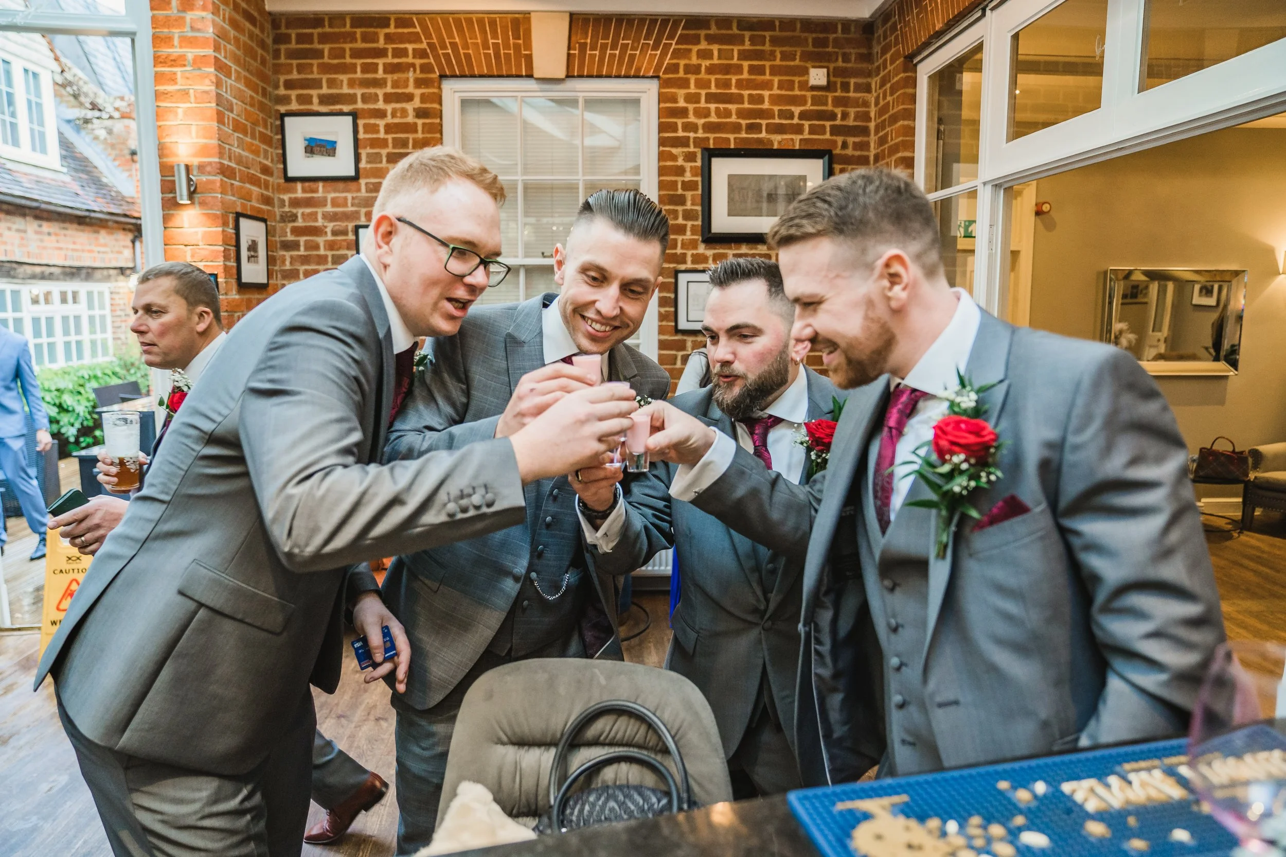 A group of five men dressed in suits, smiling and toasting with shot glasses at a social gathering indoors with brick walls and framed pictures at Warbrook House