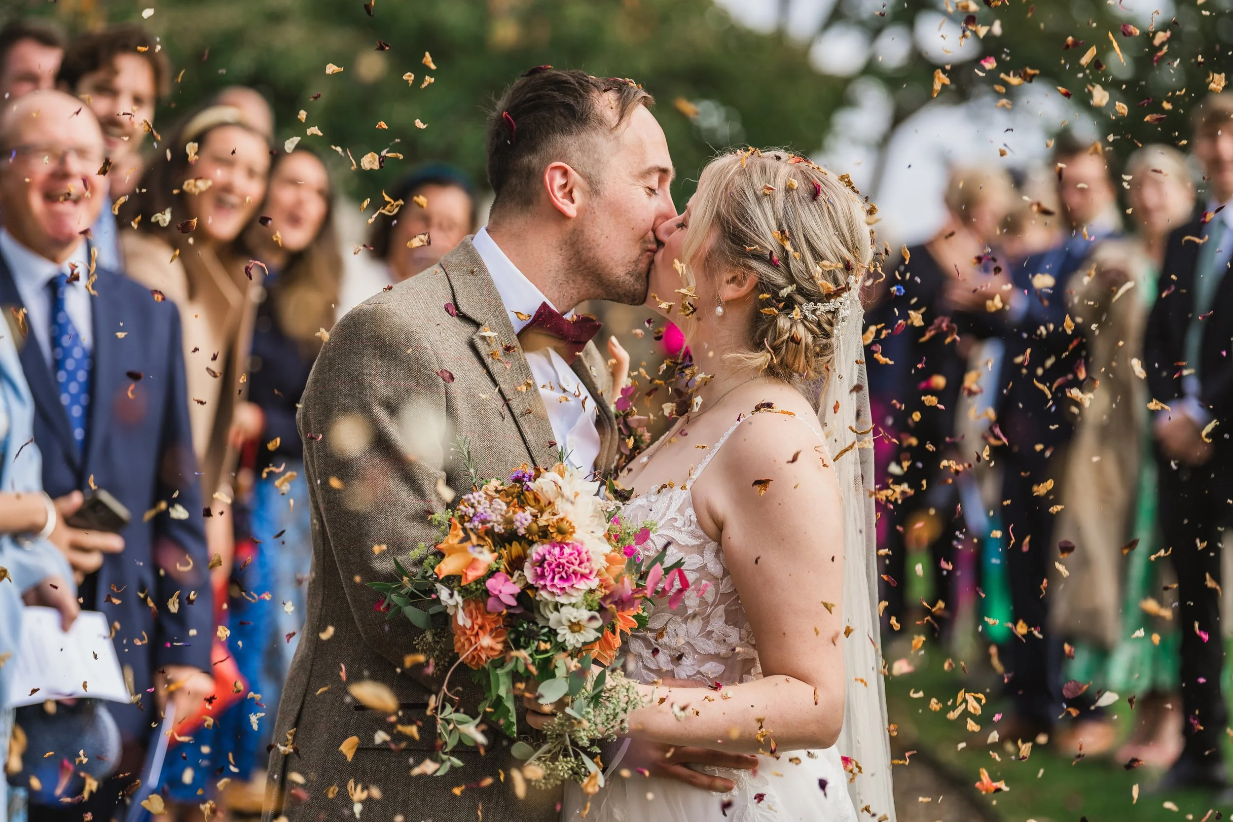 A newlywed couple kisses during outdoor wedding celebration amid falling flower petals, with guests in the background.