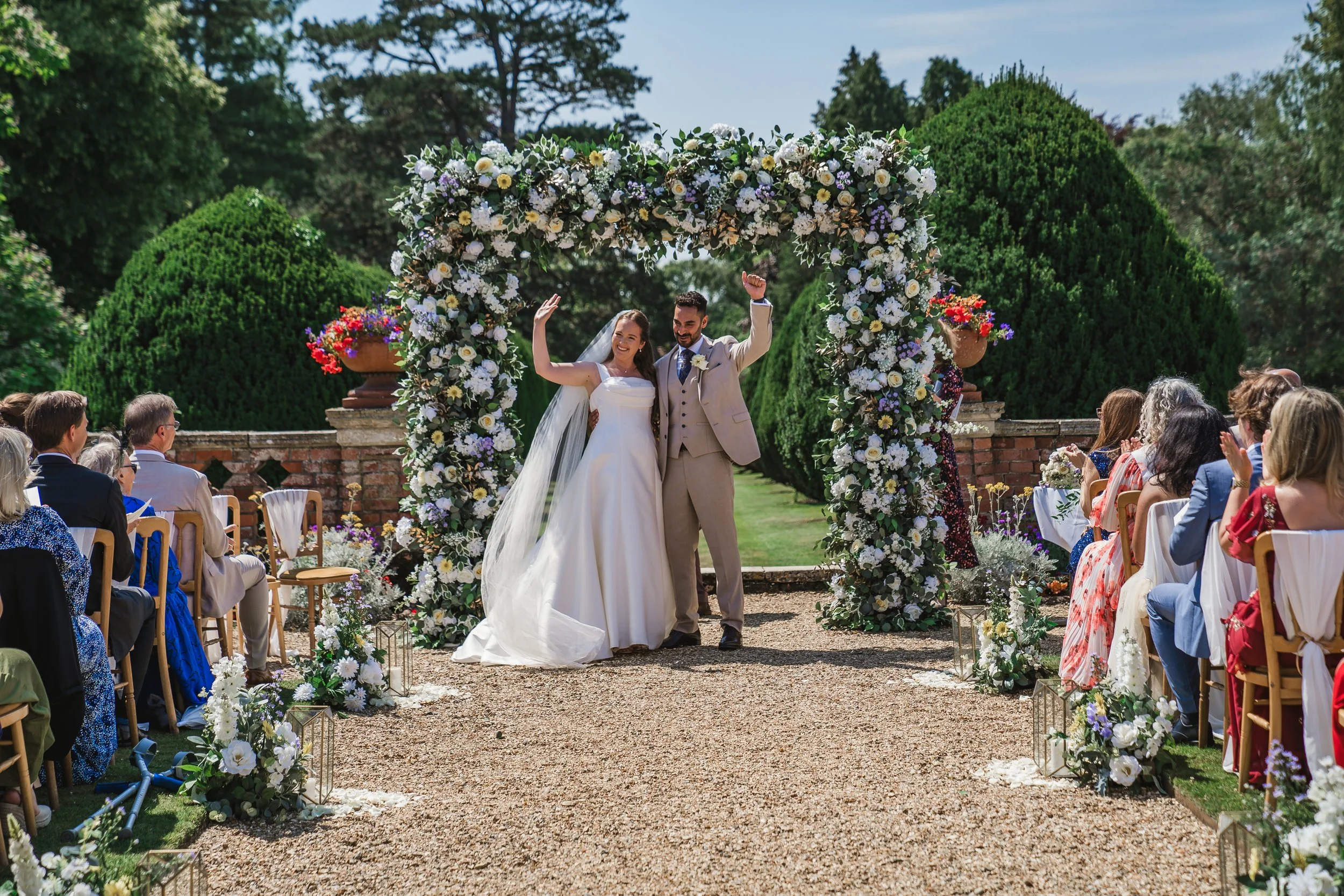 A bride and groom celebrating at their outdoor wedding ceremony under a floral arch, with guests seated on either side, in a green garden setting with trees and flowers.