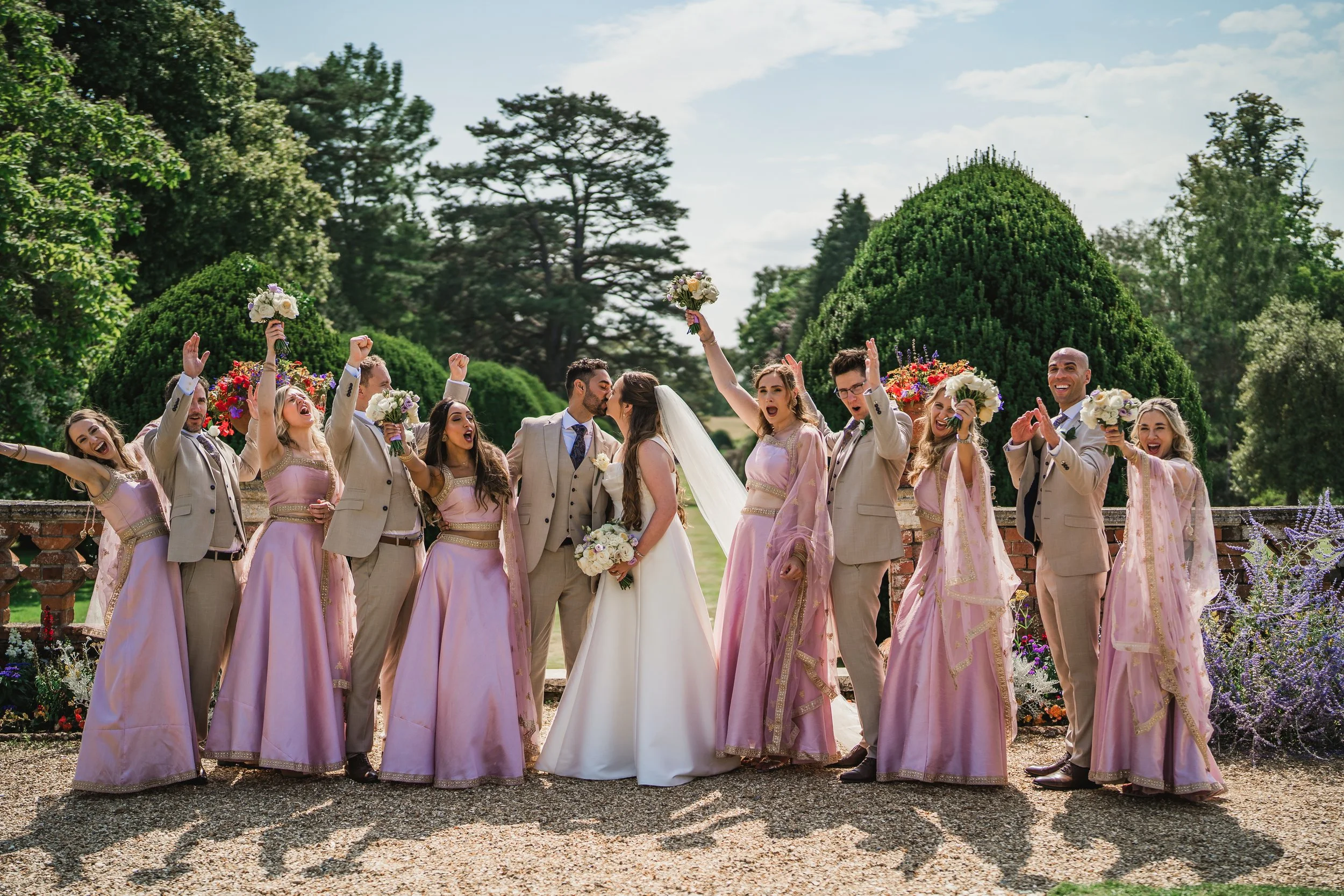 A wedding party celebrating outdoors with lush green trees and a blue sky in the background. The bride and groom kiss in the center, surrounded by bridesmaids and groomsmen happy, some holding bouquets and raising their fists in celebration.