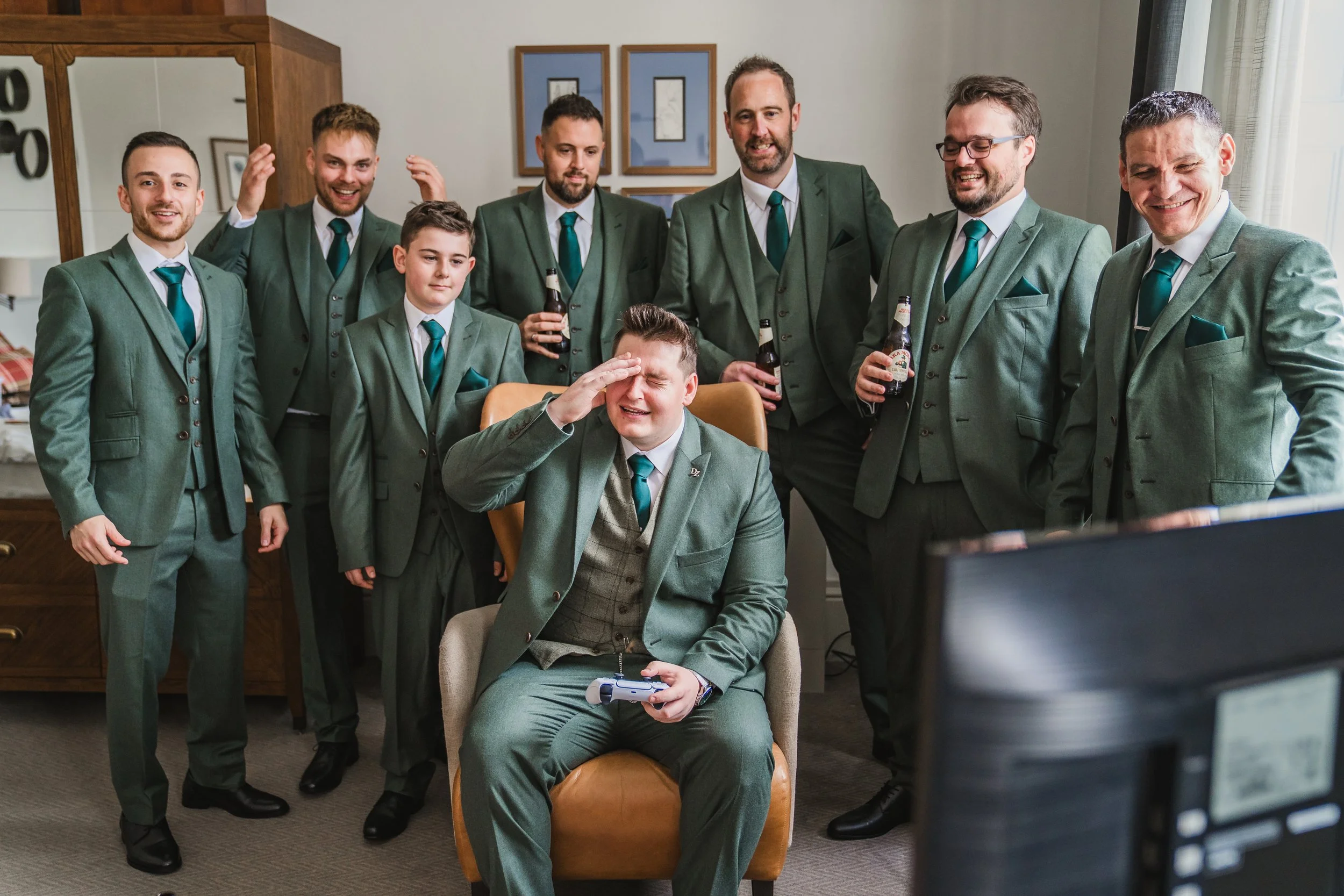 Group of nine men wearing matching green suits and teal ties, gathered in a room, with one seated in front, saluting a gaming controller, while others are standing around, holding drinks and smiling.
