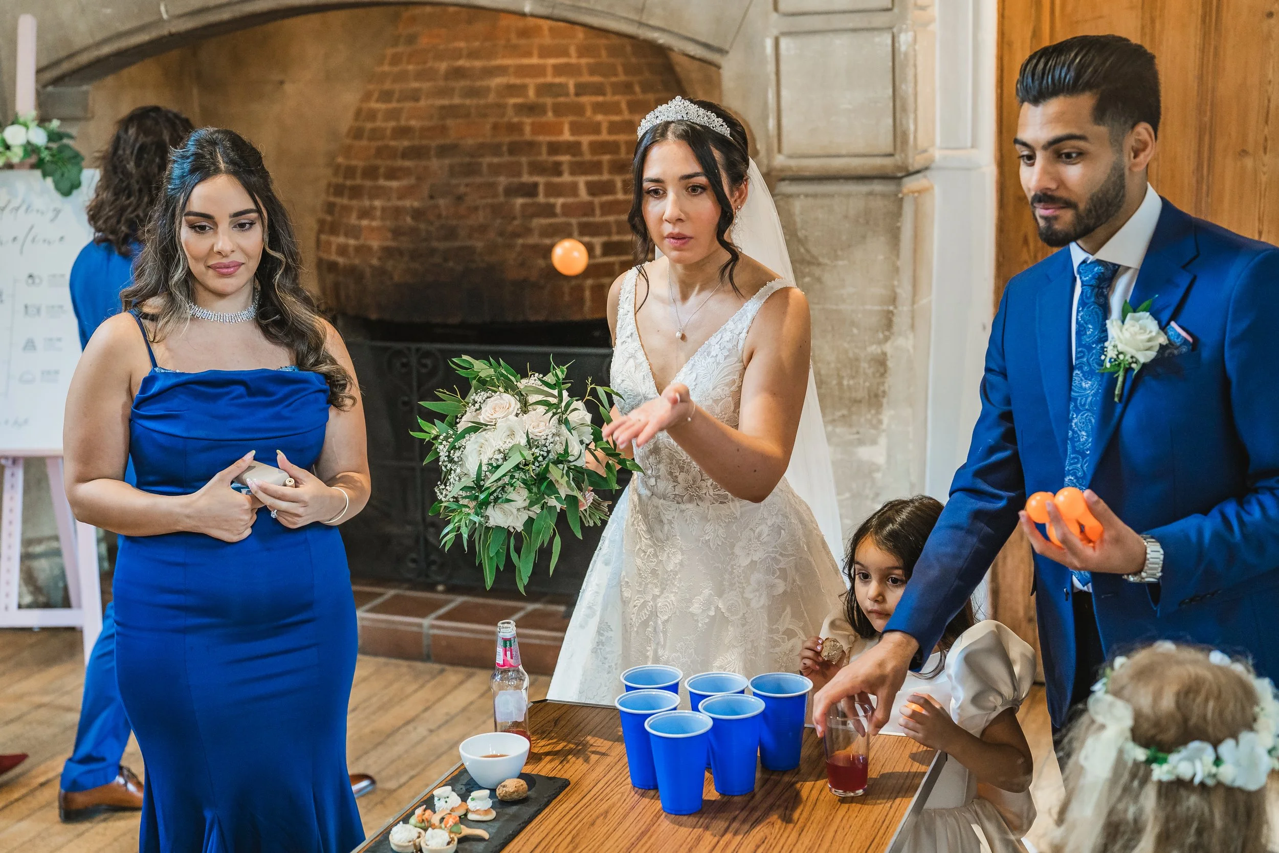 A wedding reception scene with a bride holding a bouquet of flowers, a woman in a blue dress, a man in a navy suit, and children playing drinking games at a table with blue cups and snacks in a room with brick and wooden walls.