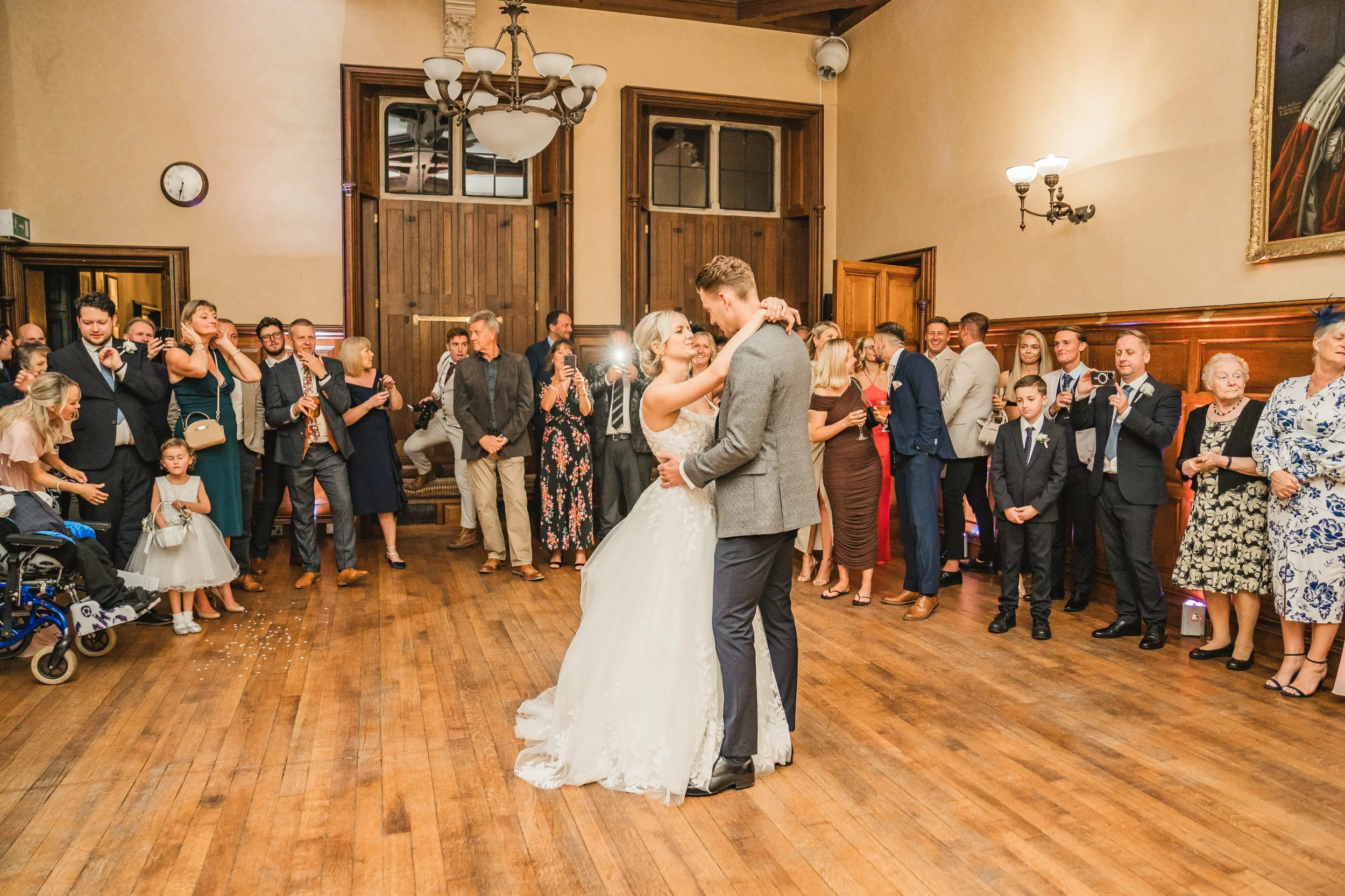 A bride and groom are dancing in the center of a room with wooden floors, surrounded by guests watching and taking photos. The bride is wearing a white wedding gown and the groom a gray suit. The room features wood-paneled walls and vintage lighting 