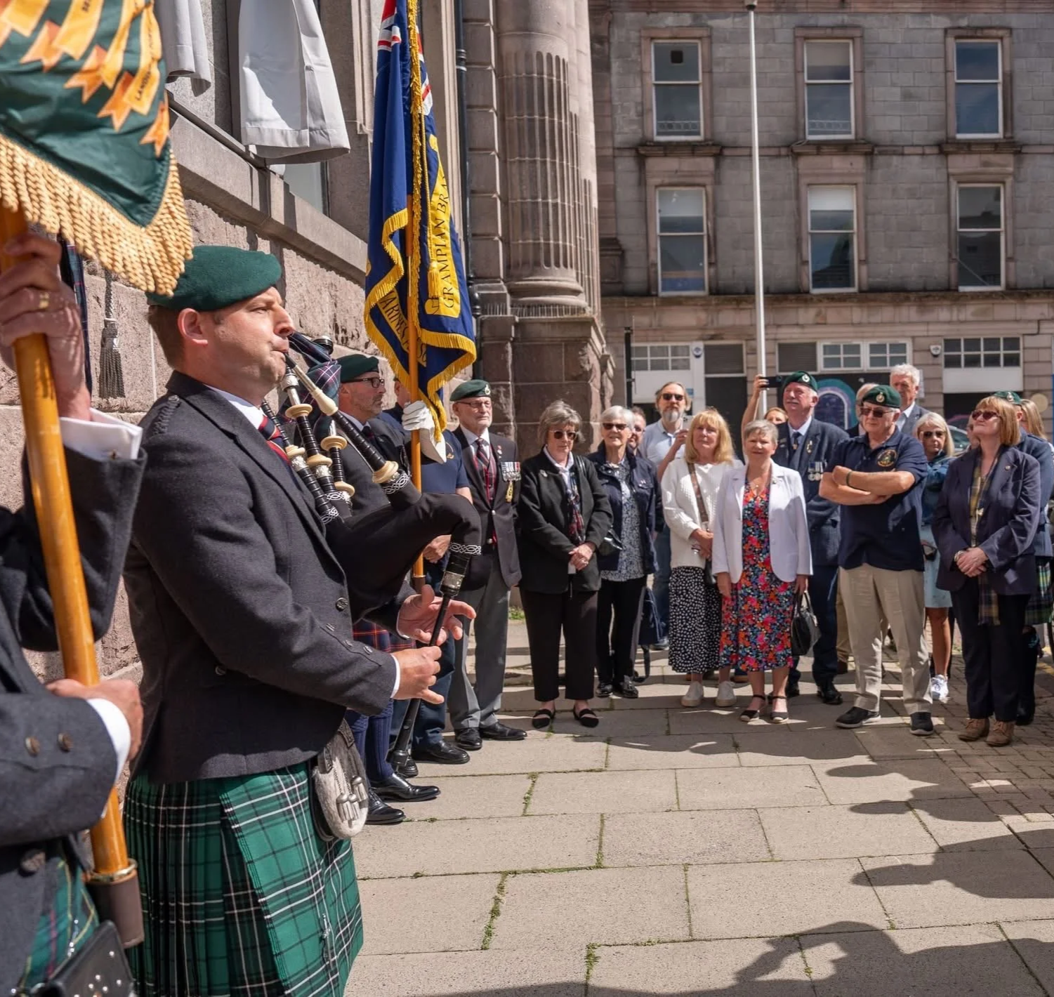 A group of people gathered outside a building, some wearing military or veteran uniforms, and some holding flags. One man in the foreground dressed in traditional Scottish attire is playing the bagpipes. It appears to be a memorial or commemorative event.