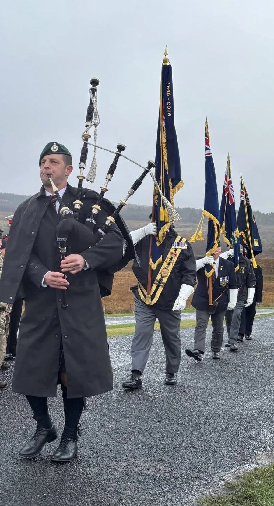 A group of veterans or military personnel marching with flags, including the Union Jack, during an outdoor ceremony or parade, with a cloudy sky and open fields in the background.