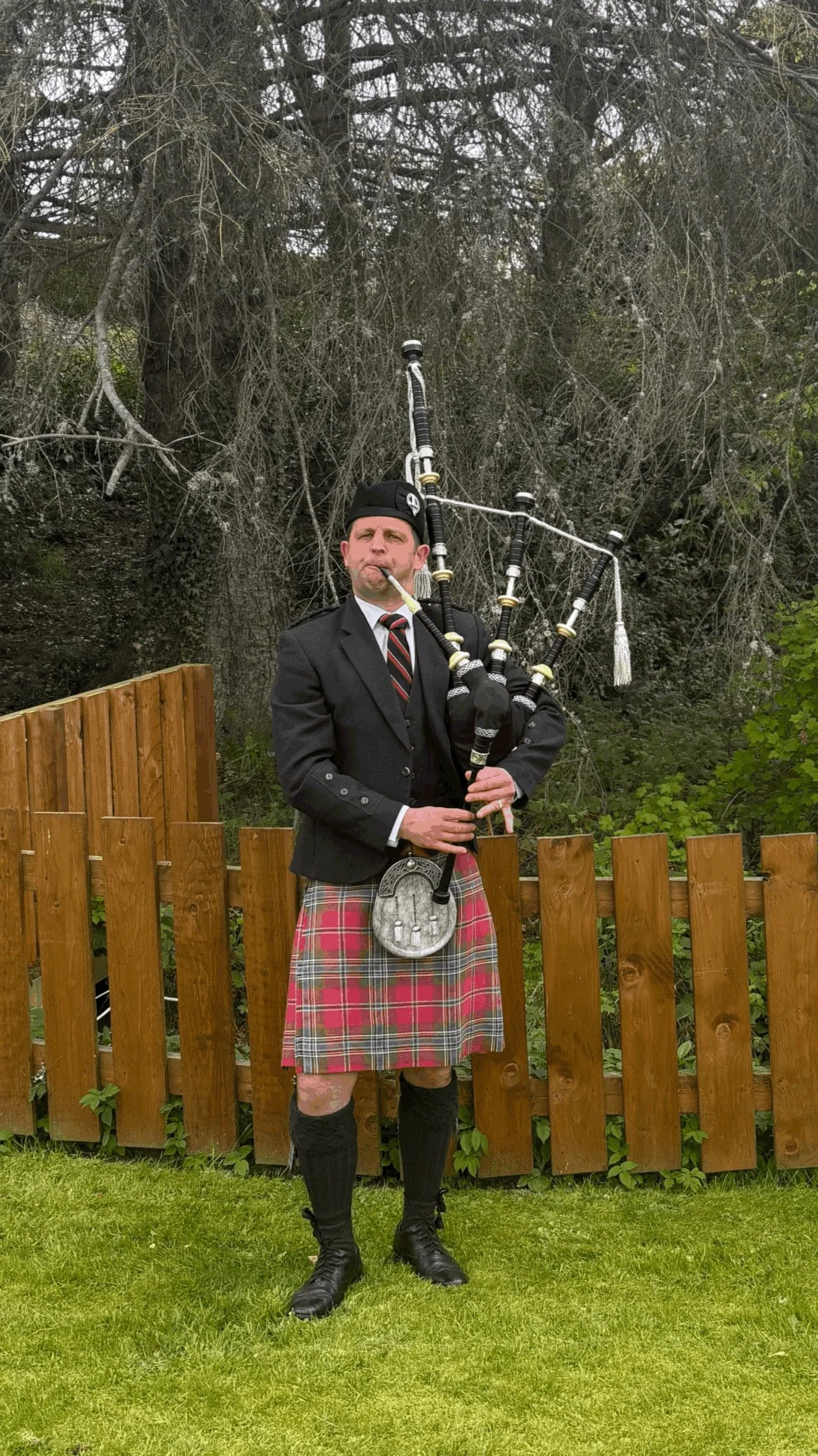 A man dressed in traditional Scottish attire playing bagpipes outdoors. He is wearing a black blazer, a red and green tartan kilt, black boots, and a black hat, standing in front of a wooden fence with trees in the background.
