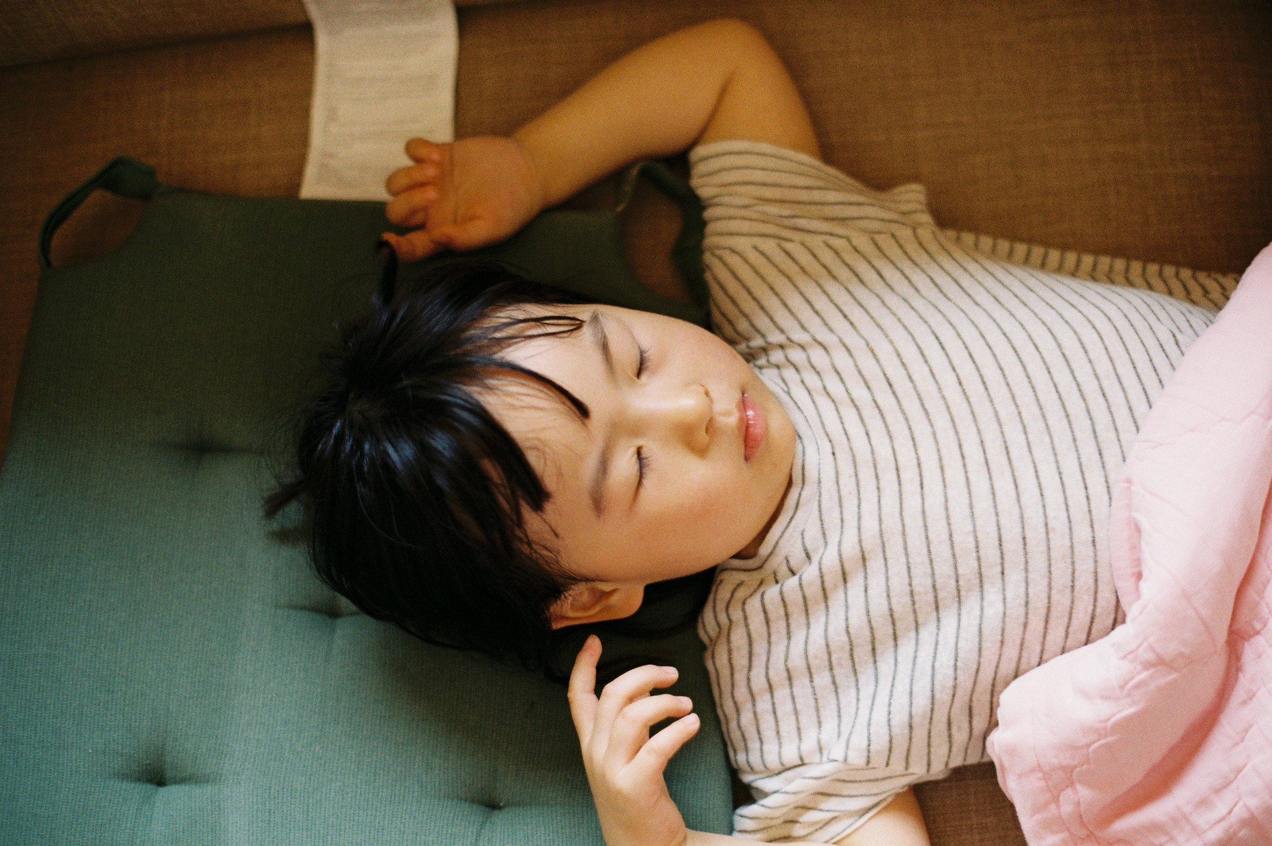 A young girl with short black hair sleeping peacefully on a green couch, with her head resting on a green pillow, wearing a striped shirt and covered with a pink blanket.