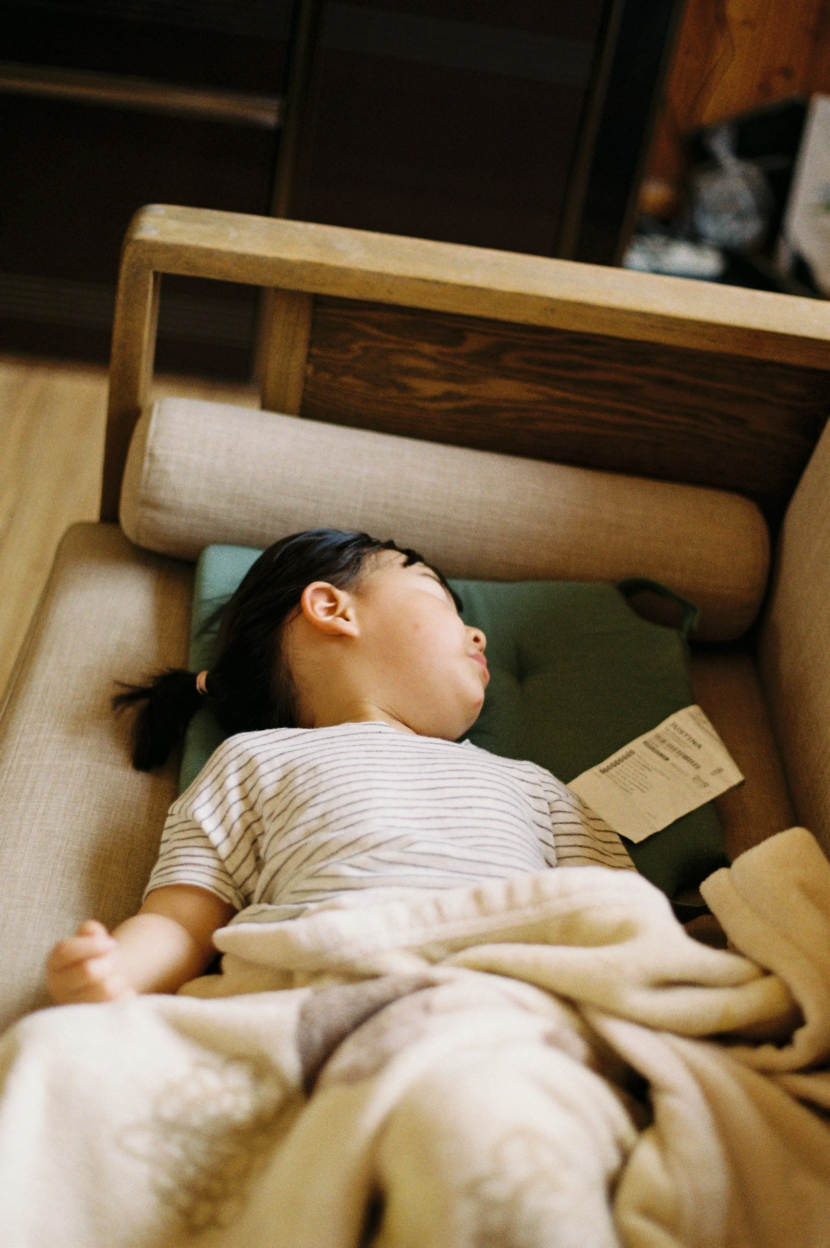 A young girl with dark hair resting on a sofa, sleeping, with a pamphlet in her hand and a cozy blanket covering her.