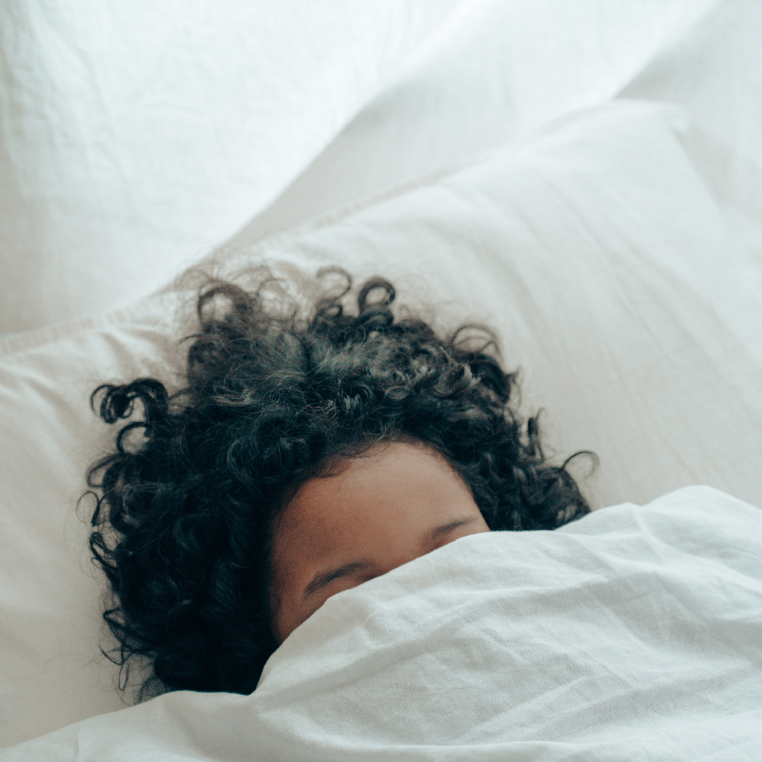 Close-up of a person with curly hair sleeping in a bed, partially covered by white sheets.