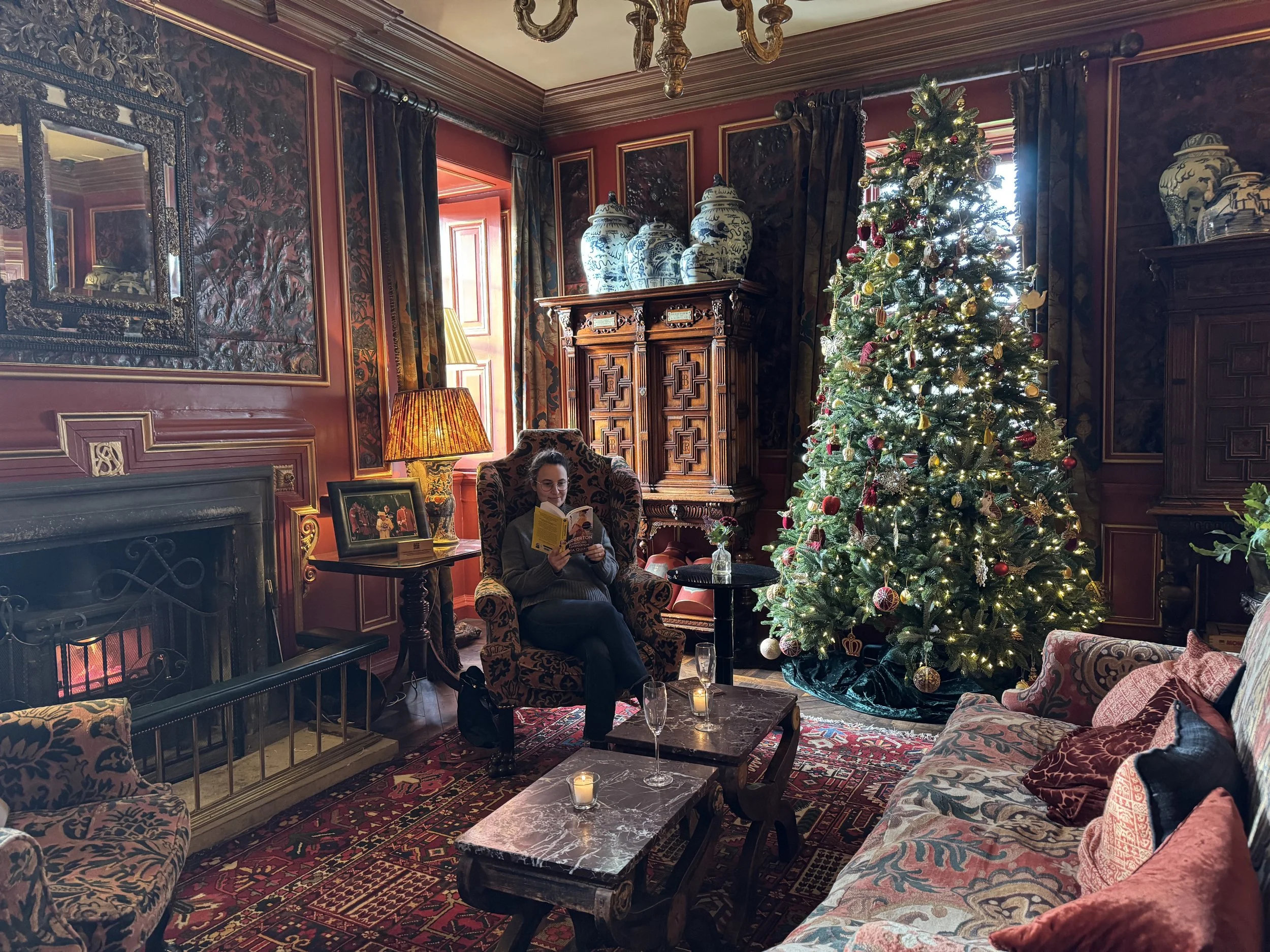 A woman sitting in an ornate armchair in a decorated living room with a large, decorated Christmas tree, antique furniture, artwork, and a fireplace.