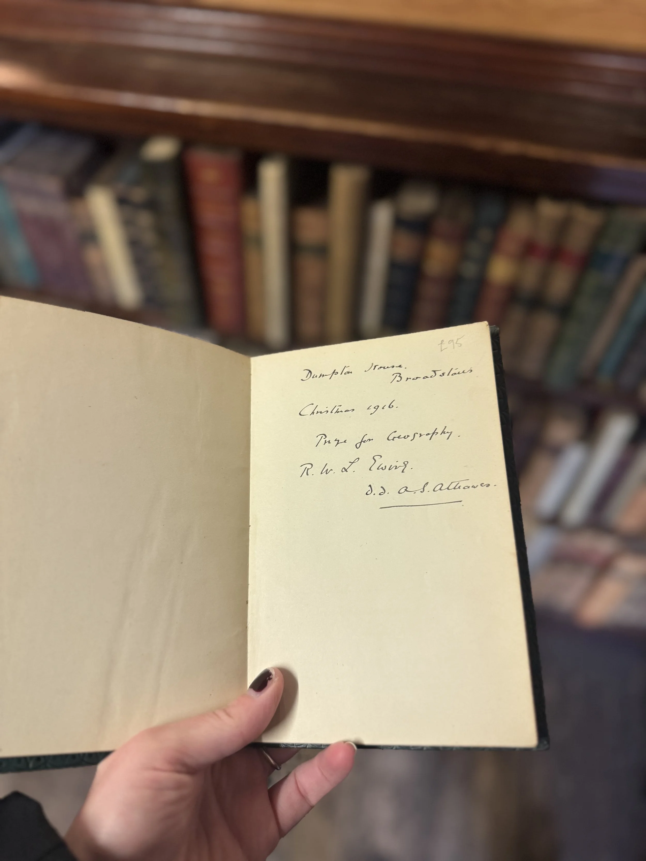 Hand holding an open notebook with handwritten text, with a bookshelf in the background.