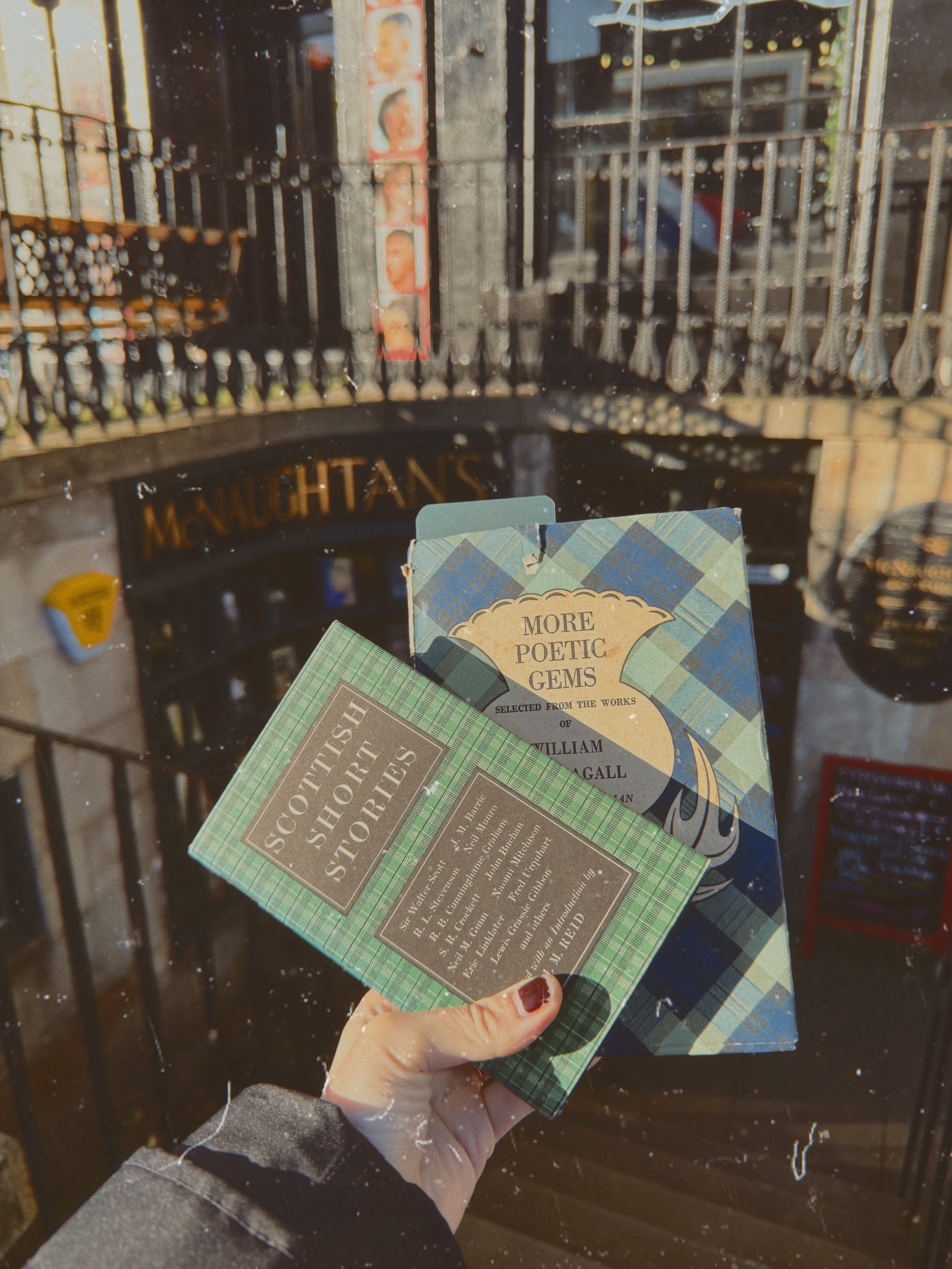 A person holding three books titled "Scottish History Stories," "More Poetic Gems," and other titles, in front of a store window with a black sign that has the word "MANHATTAN" on it.