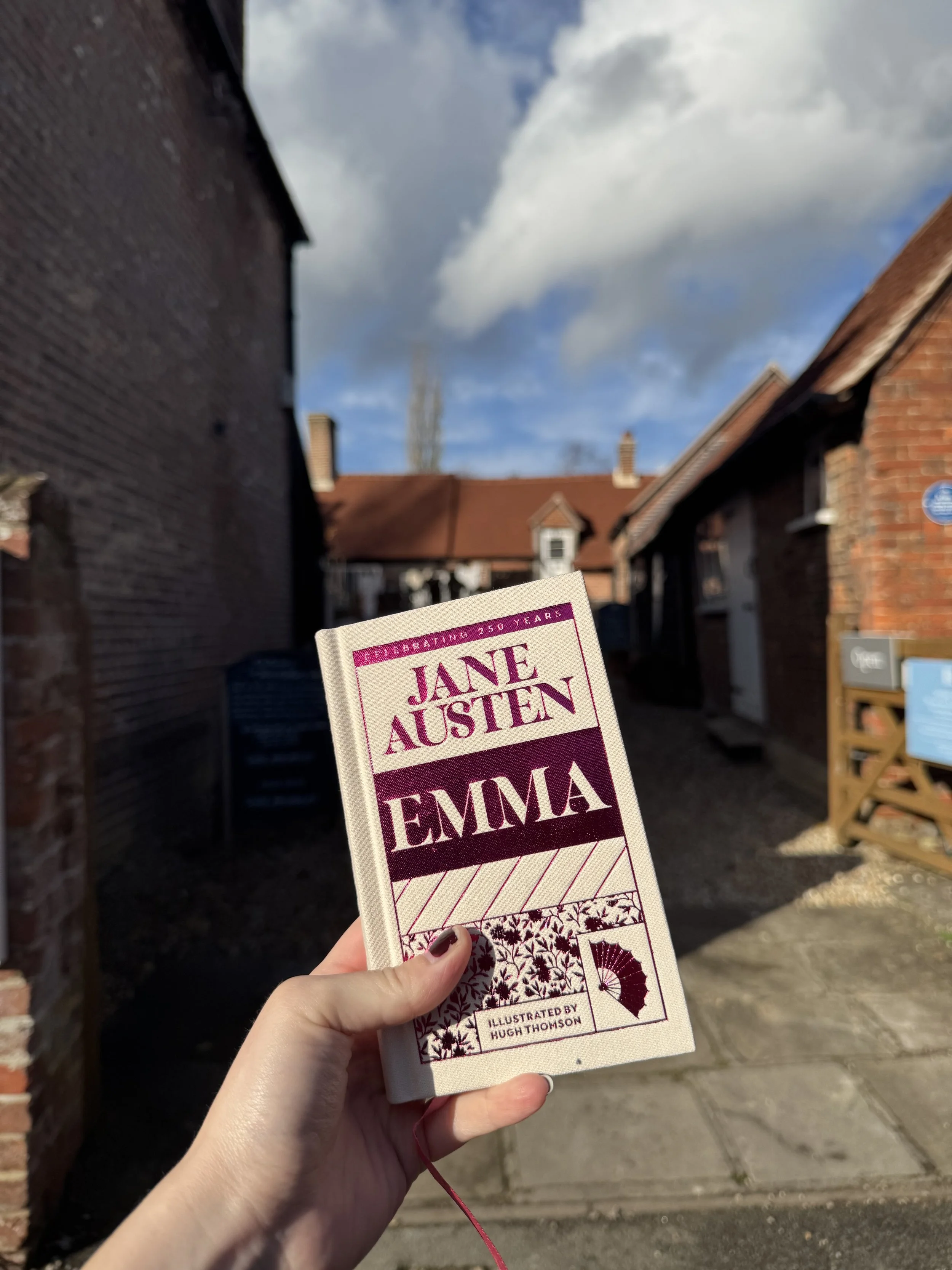 Person holding a paperback book titled 'Emma' by Jane Austen, with a background of brick buildings and a partly cloudy sky.