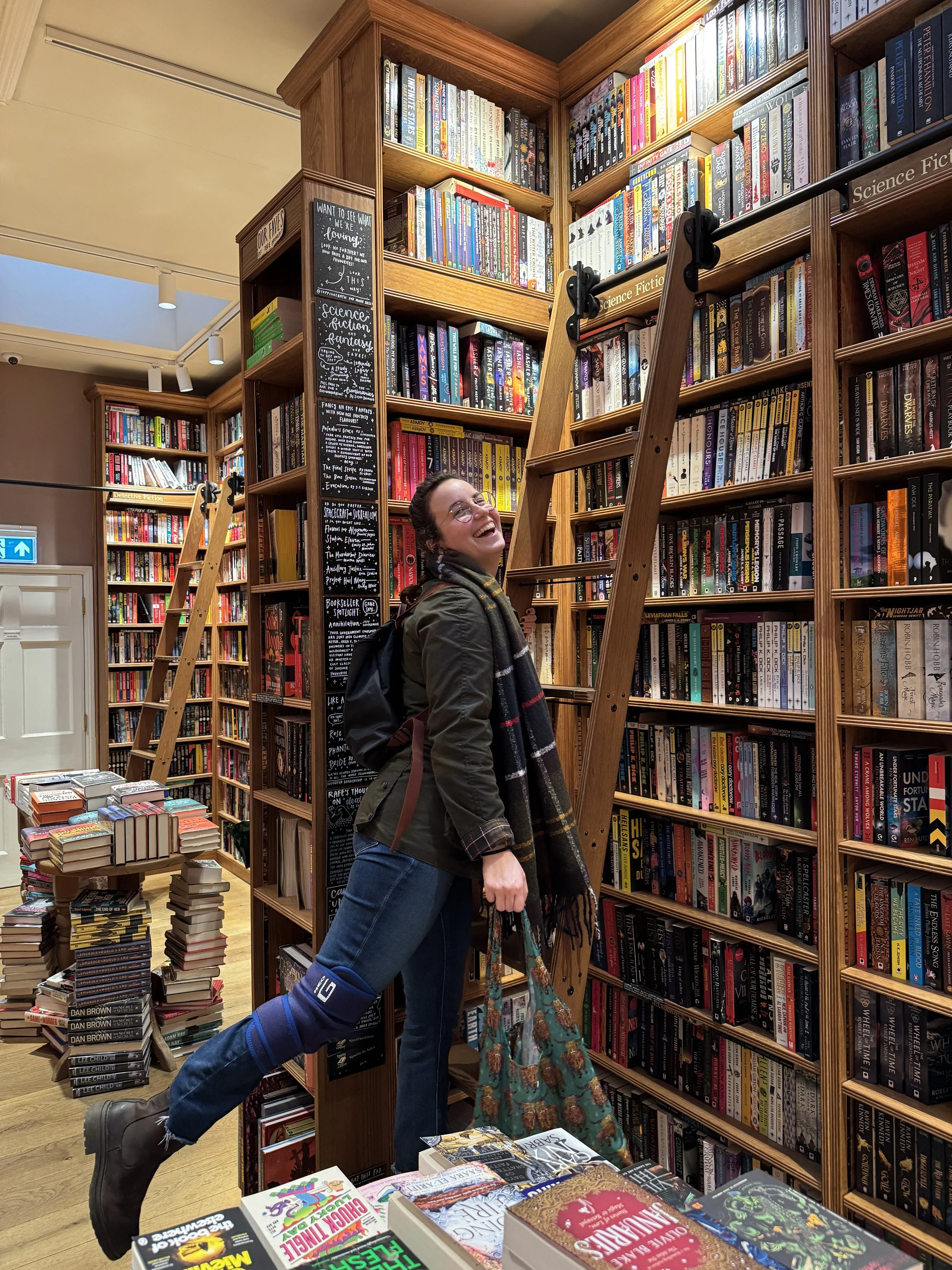 A person with glasses and dark hair smiling and laughing while standing in a bookstore aisle, surrounded by tall wooden bookshelves filled with science fiction and fantasy books. The person is wearing a dark jacket, plaid scarf, blue jeans, and brown boots, and is leaning on a ladder.