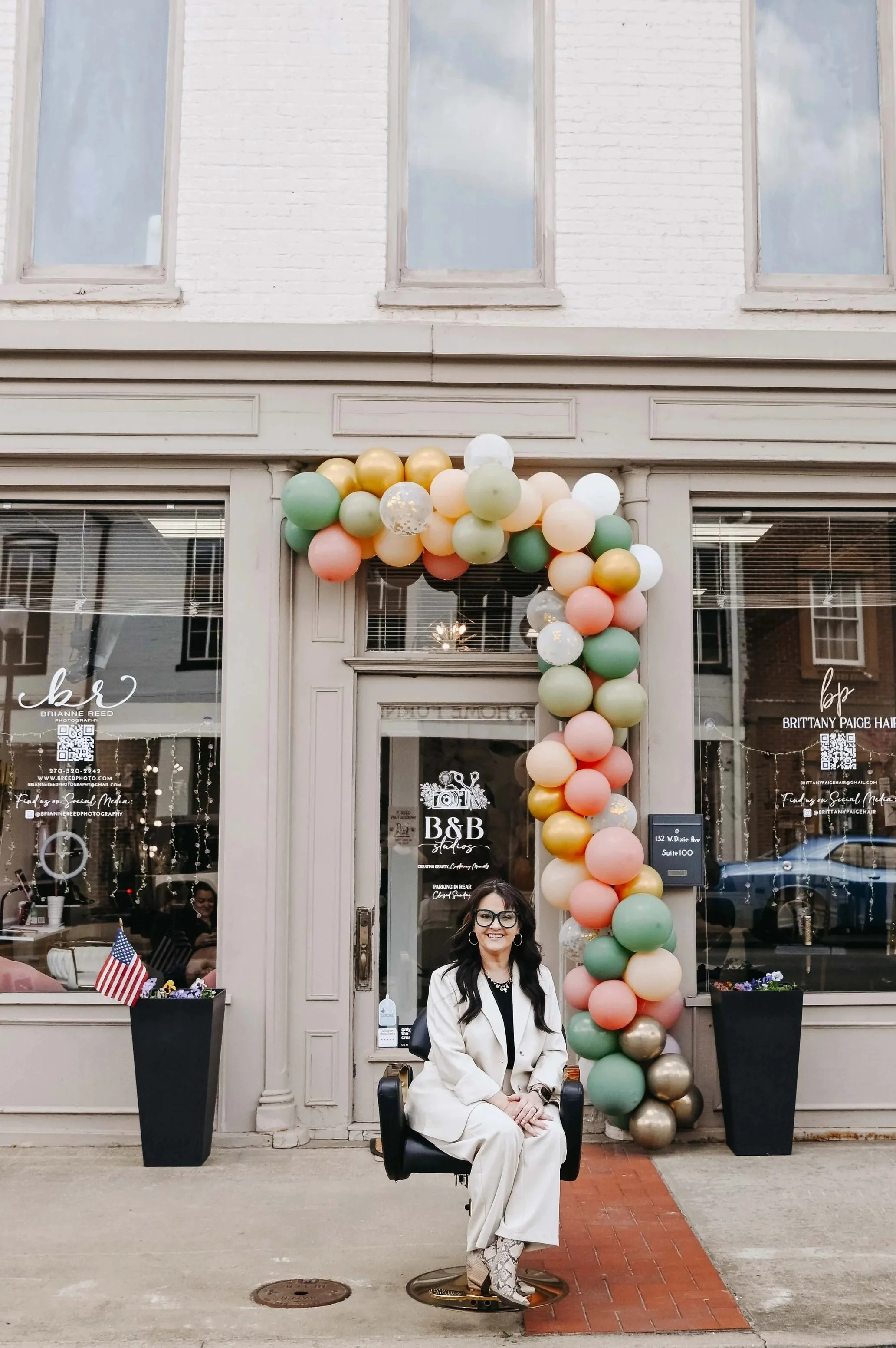 A woman sitting on a black chair in front of a storefront decorated with balloons. The store has large windows with blinds and signages, and two tall black planters with American flags on either side of the entrance.