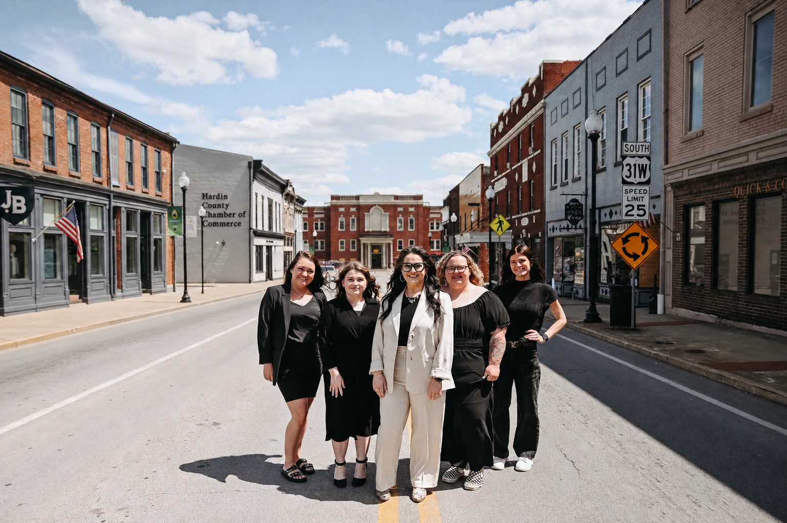 Five women standing together on a city street, smiling. Buildings and street signs in the background, clear sky above.
