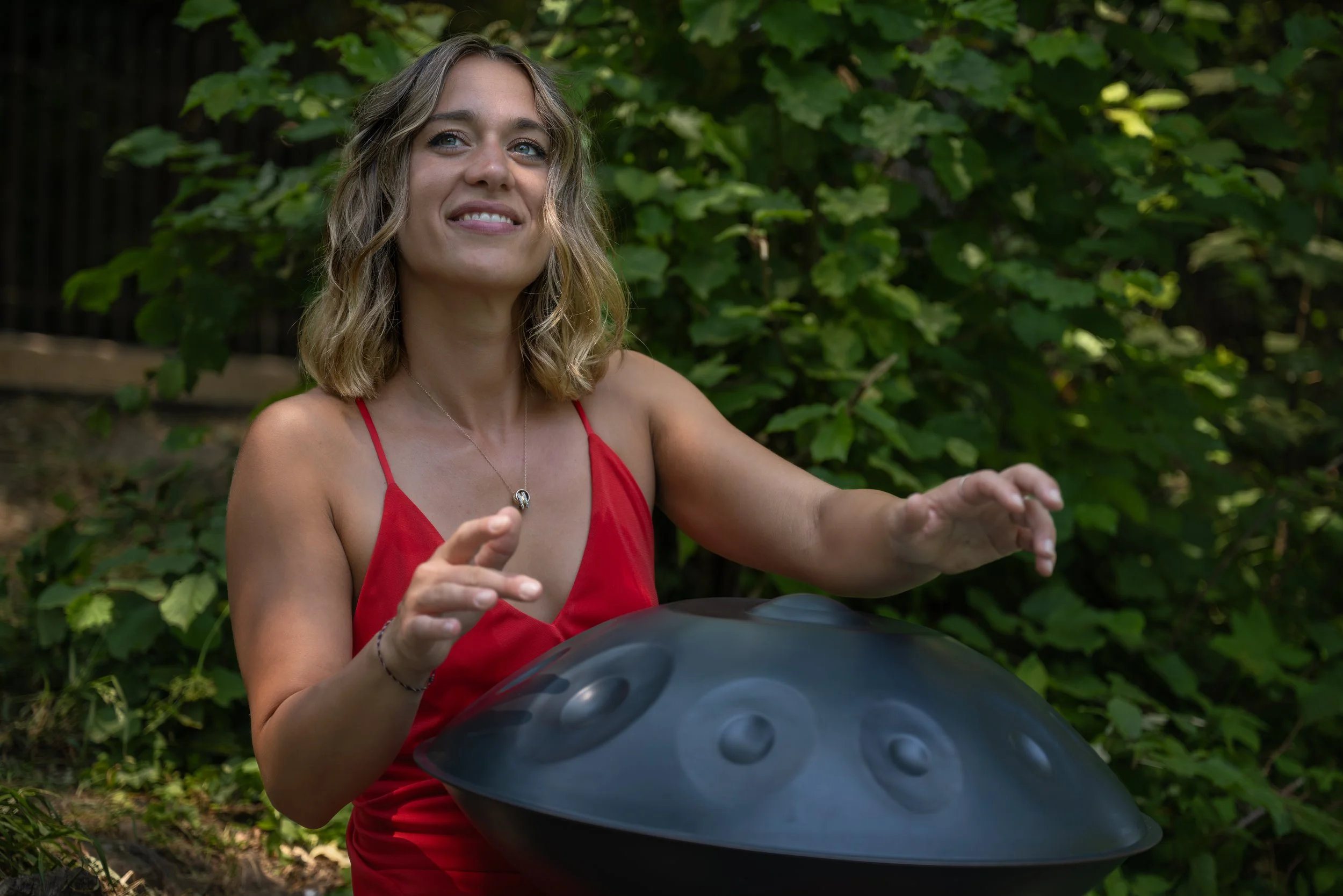 Berni Ditroi in a red dress smiling and playing handpan outdoors, contact for bookings and enquiries.