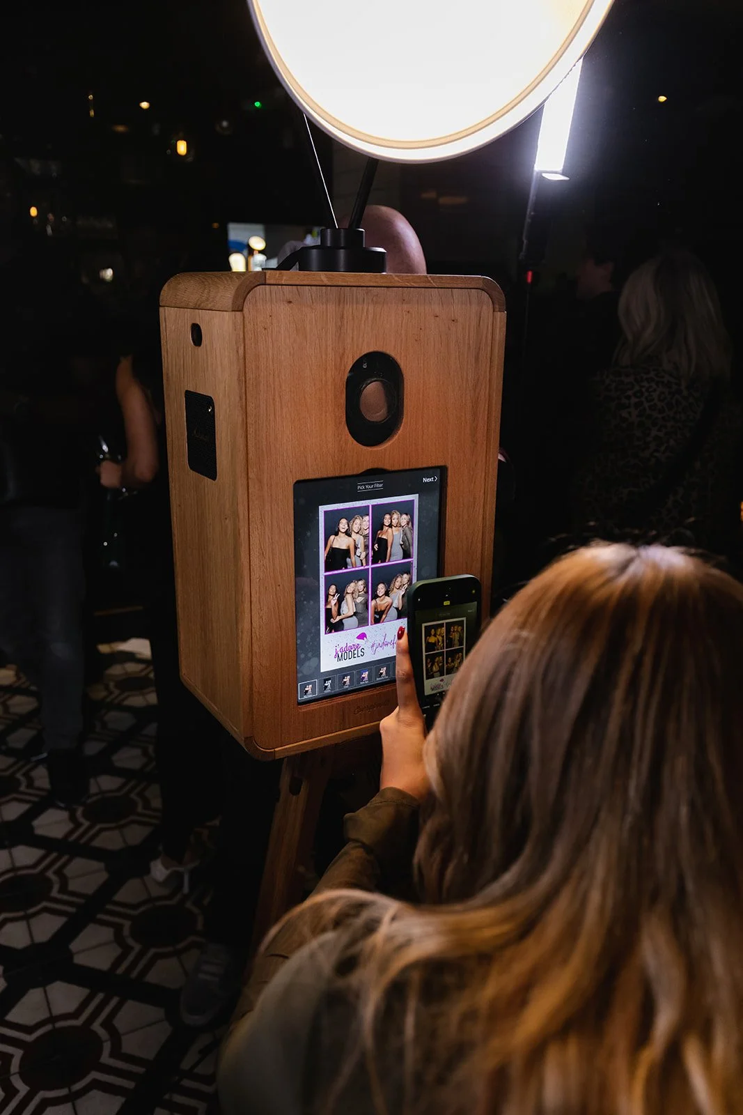A woman with long, wavy blonde hair is using a smartphone to select photos on a touchscreen photo booth at an event. The photo booth has a wooden exterior and a large circular light on top. Other guests are visible in the background.