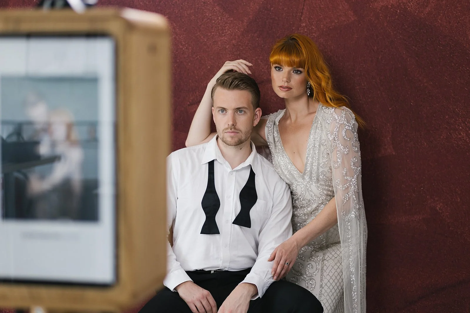 A man and woman seated against a red wall are being photographed. The man is wearing a white shirt with a black tie, and the woman has red hair and is dressed in a silver embellished gown. The woman is leaning close to the man with her arm around his shoulders, and they both have serious expressions. The photo camera is visible in the foreground.