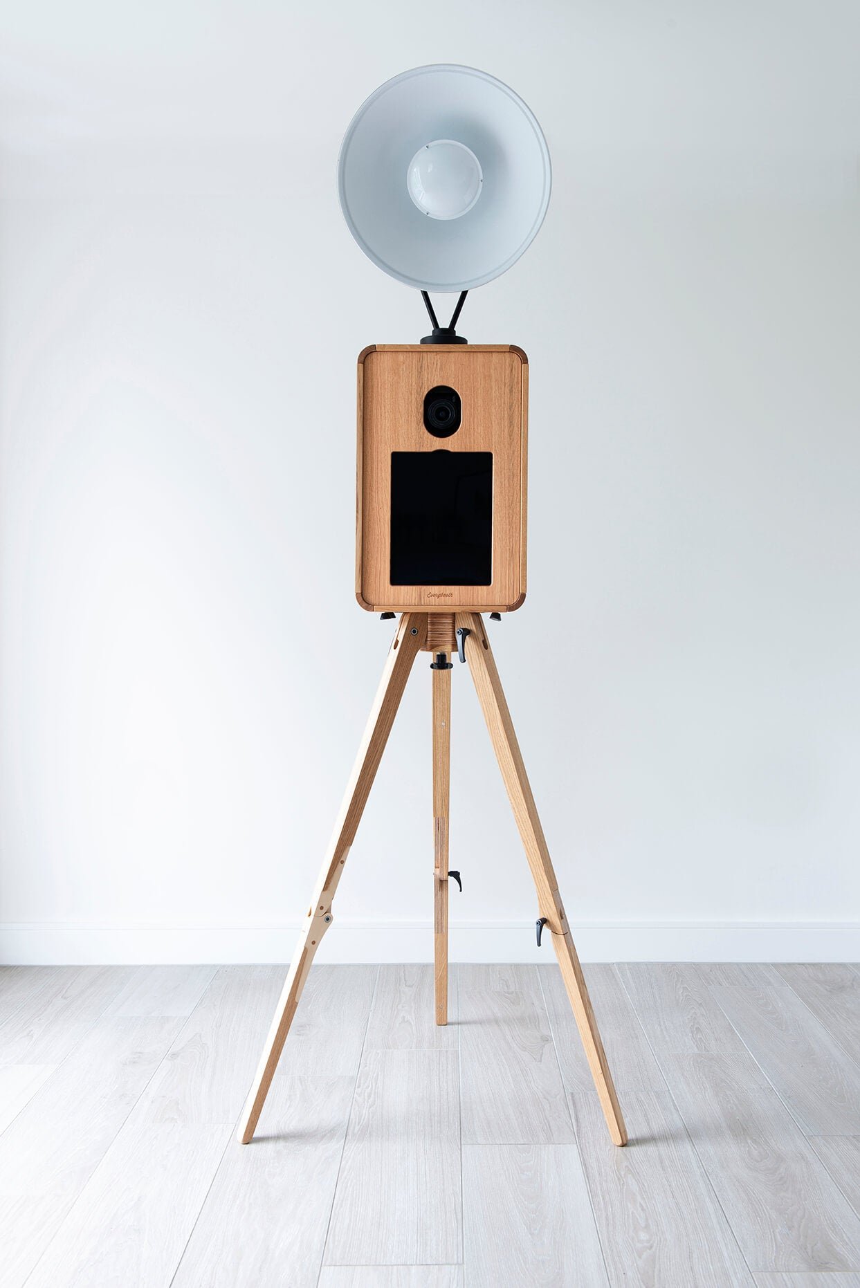 A wooden tripod stand with a large white light fixture and a camera on top, positioned against a blank white wall.