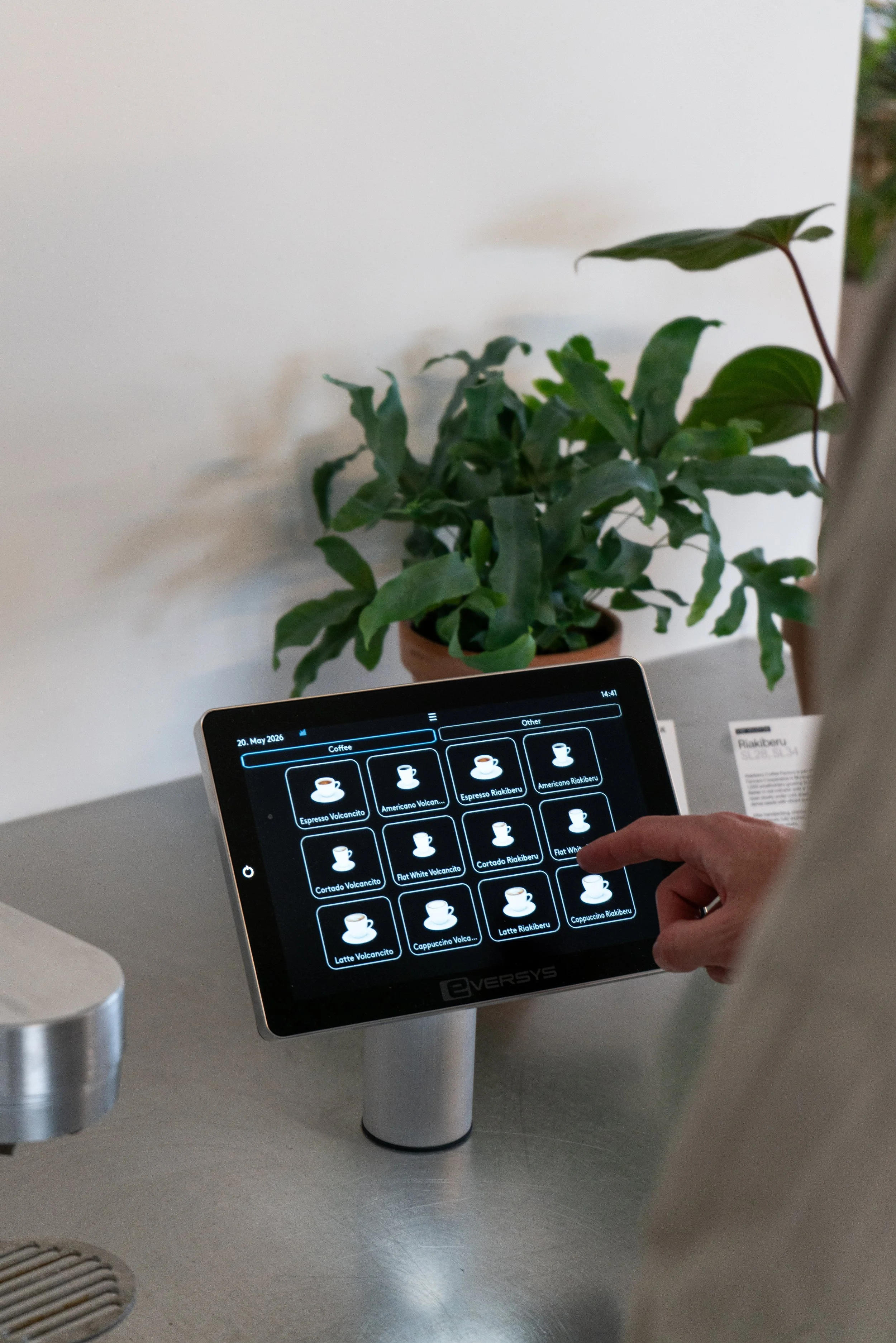 A person is using a touchscreen coffee ordering kiosk. The screen displays various coffee options with icons and names, including espresso, americano, cappuccino, latte, and cortado, with the date "20. May 2026" at the top. There is a potted plant with green leaves in the background, and part of a stainless steel countertop is visible.