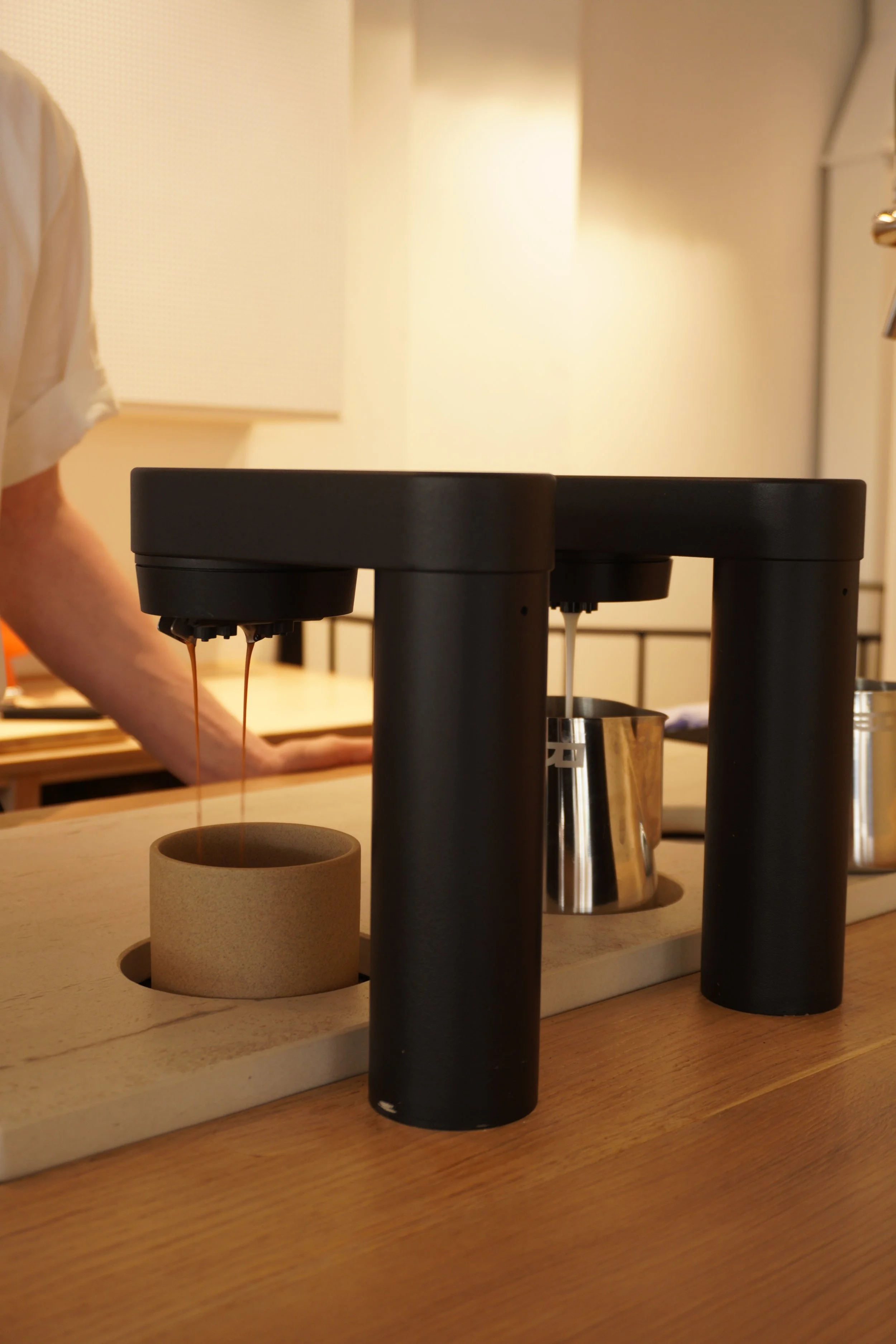 Coffee pour-over setup with two black vertical cylinders, a metal gooseneck kettle, a beige cylinder-shaped cup, and a rectangular wooden board on a wooden table.