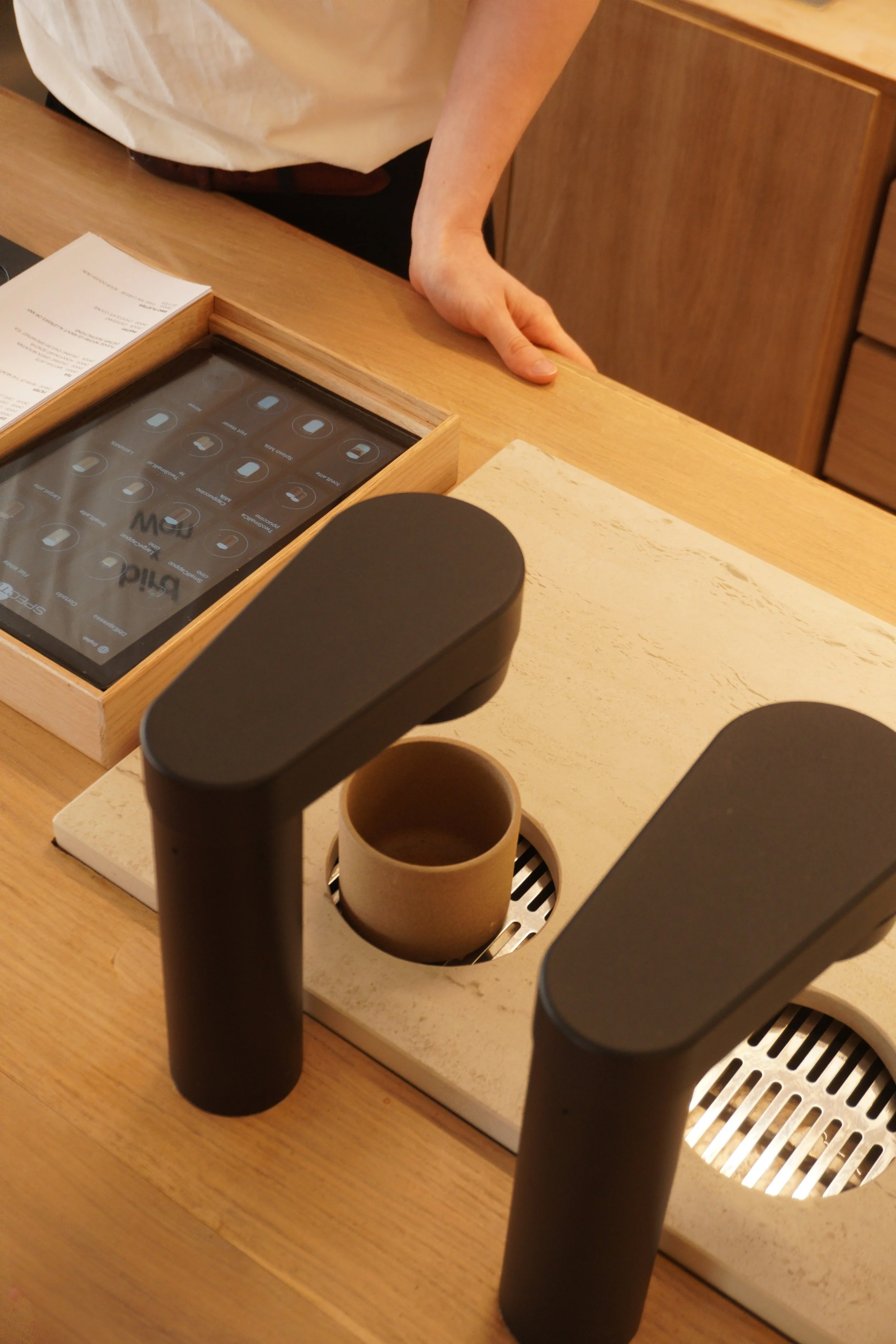 Close-up of a person making coffee at a coffee shop counter, with a latte art menu tablet, coffee cups, and a drip coffee station visible.