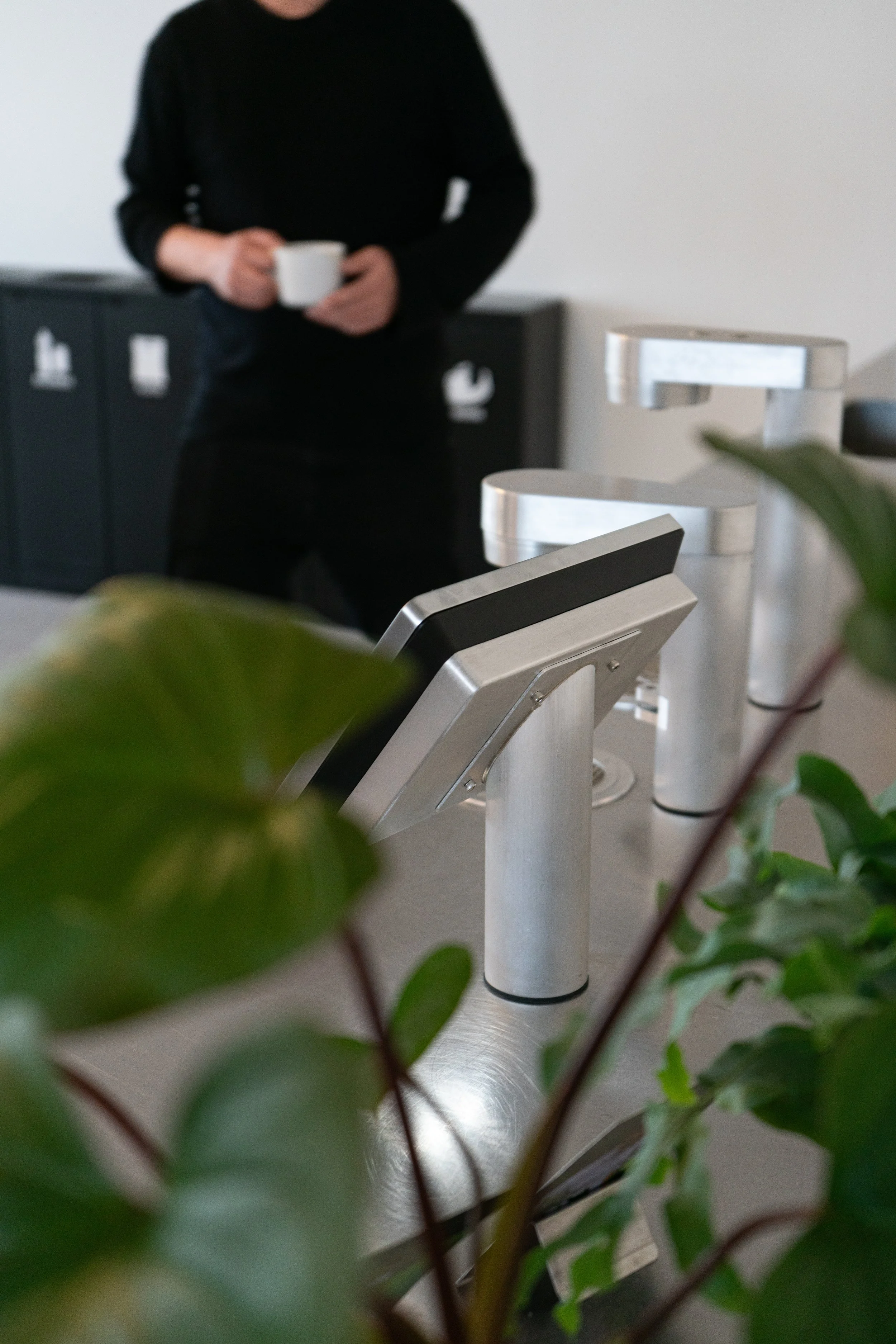 Person in black shirt holding a white coffee mug in a modern office or cafe setting, with green plant leaves in the foreground.