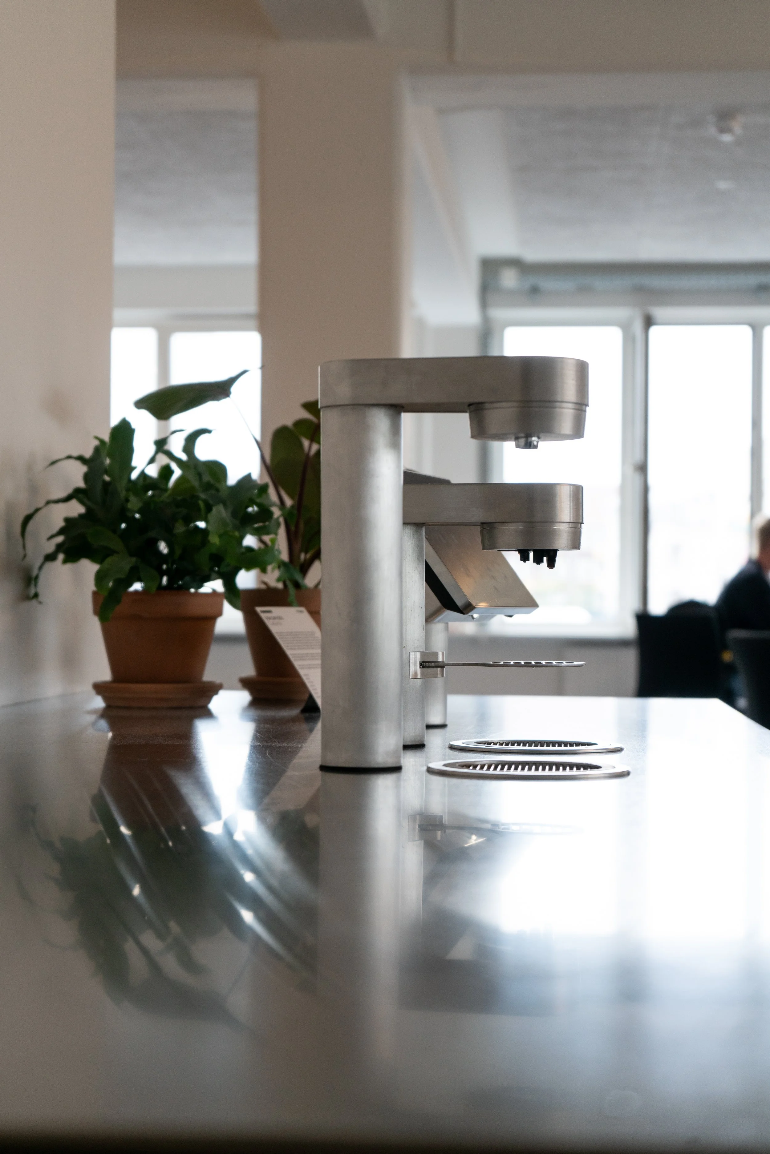 A stainless steel coffee machine placed on a countertop with two potted plants next to it, in a bright room with large windows in the background.