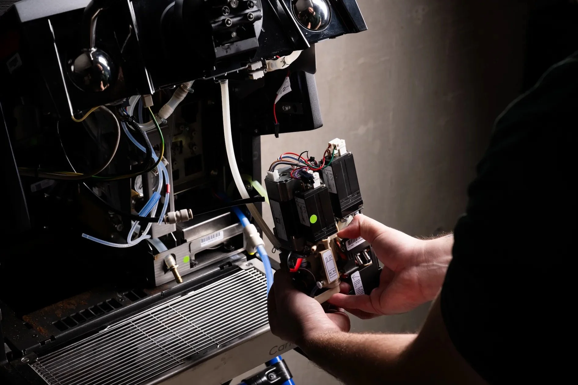 A person working with electronic components, holding a black device with wires connected to it, inside a machine with various cables and parts.