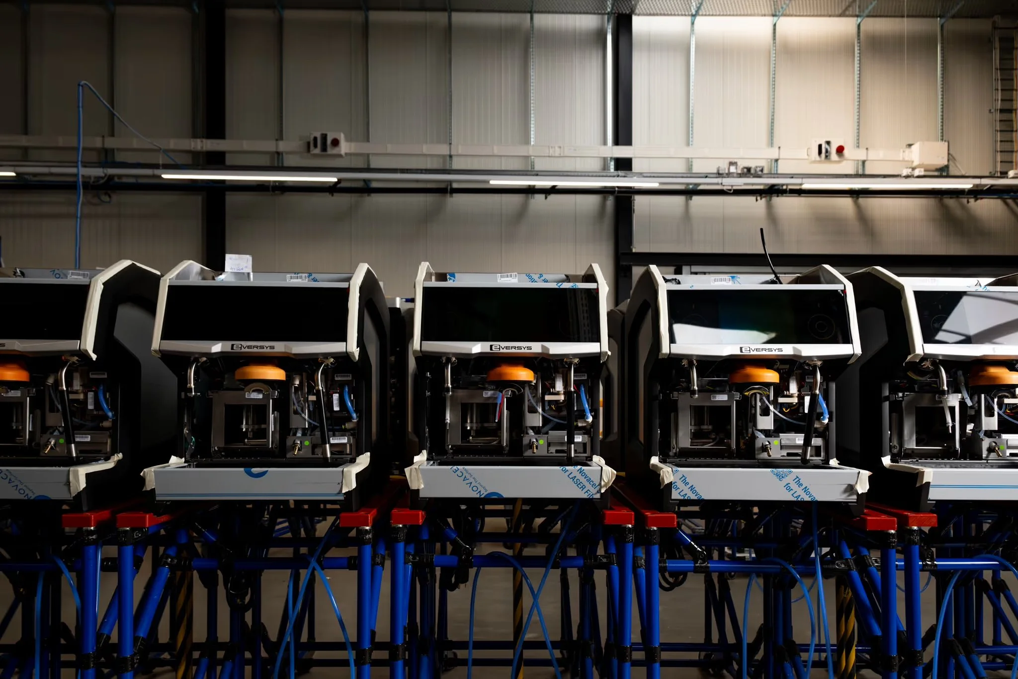 Industrial equipment with multiple robotic or electronic units connected by blue and black cables, mounted on metal supports, in a factory or warehouse setting.