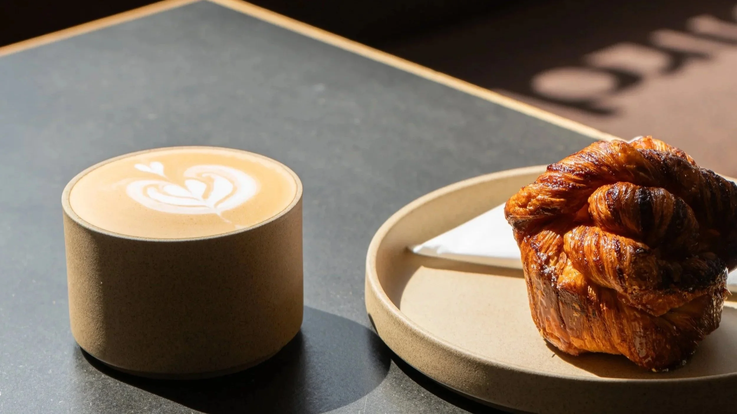 A beige cup of latte with heart-shaped foam art, placed next to a plate with a chocolate croissant on a dark surface.
