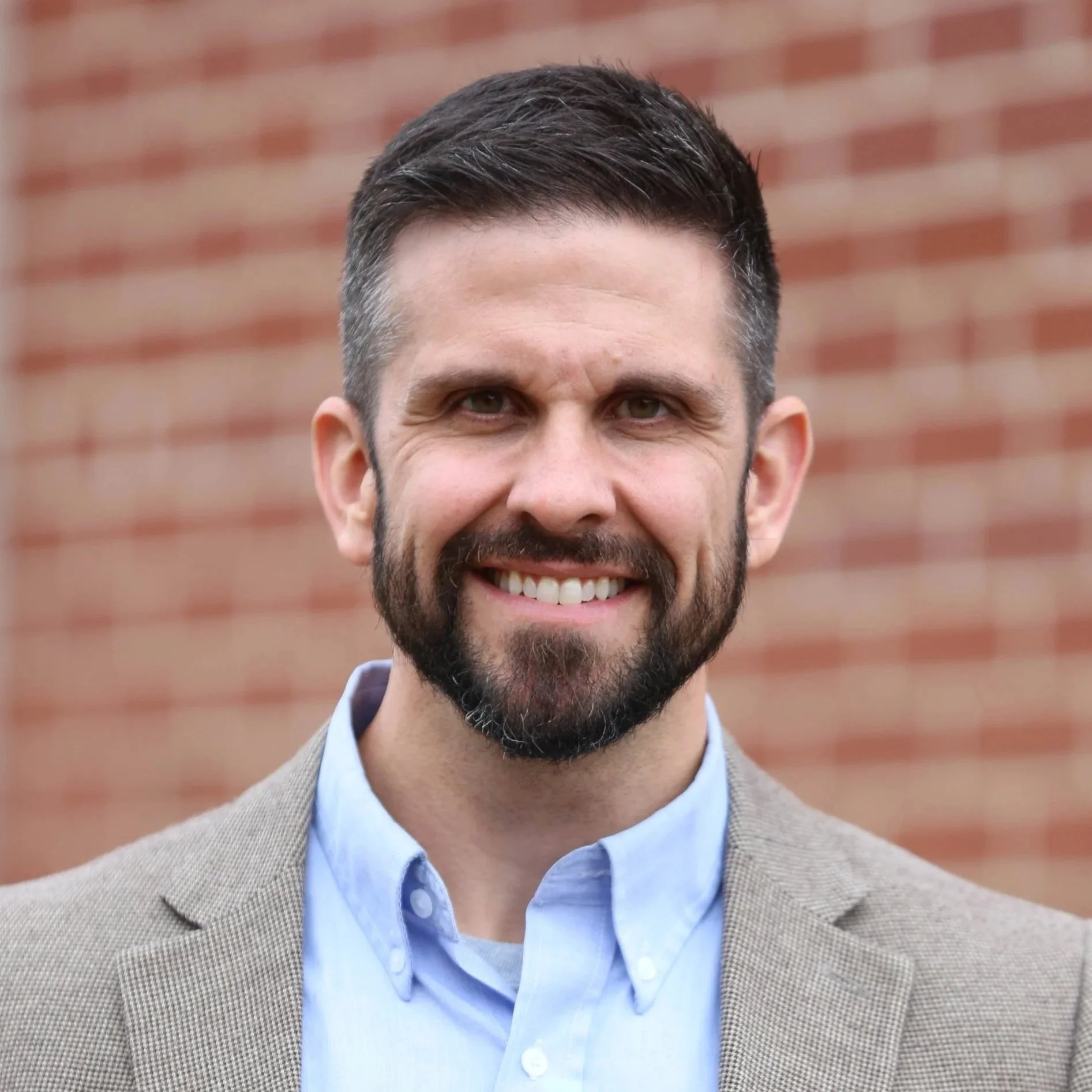 Close-up of smiling man with short dark hair, beard, and mustache, wearing a beige blazer over a light blue button-up shirt, standing against a brick wall background.
