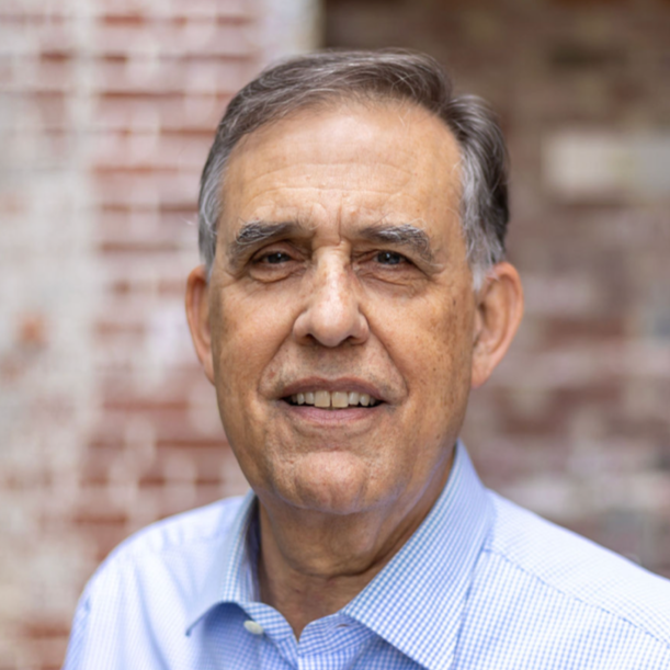 Close-up of a smiling middle-aged man with gray hair wearing a blue dress shirt, standing outdoors with a blurred brick wall background.