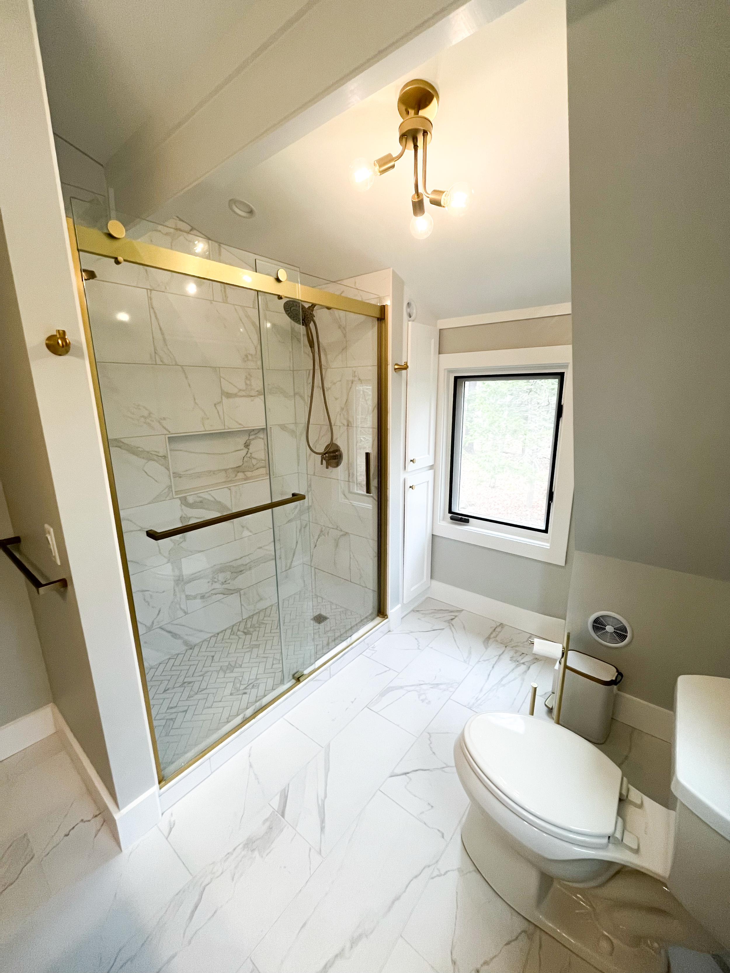Modern bathroom with white marble floor and shower with glass door and gold hardware, a window with a view of trees, and a toilet.