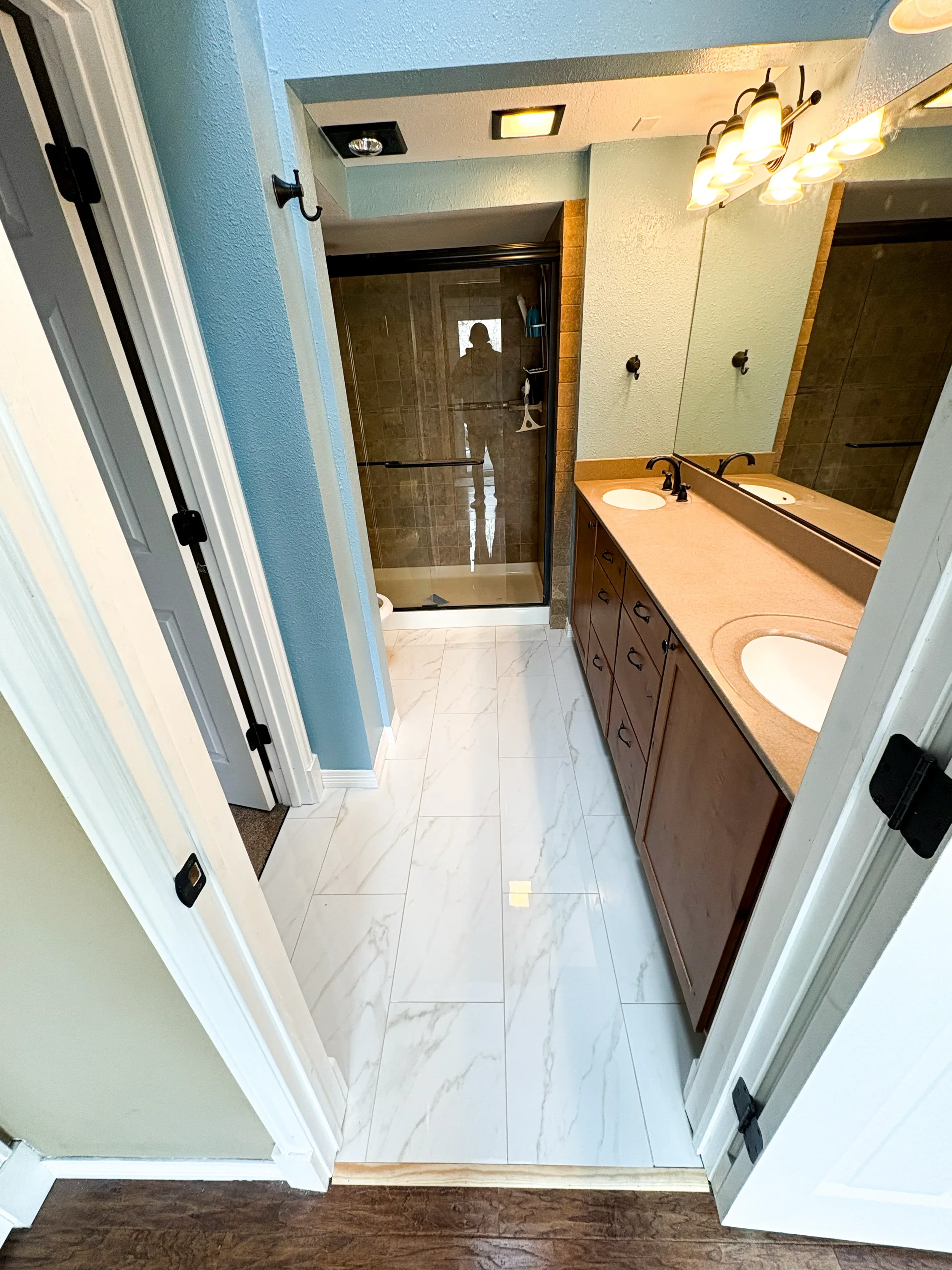 View of a bathroom with a double sink vanity, large mirror, and a glass-enclosed shower with brown tiles, white marble tile flooring, and blue-green walls.