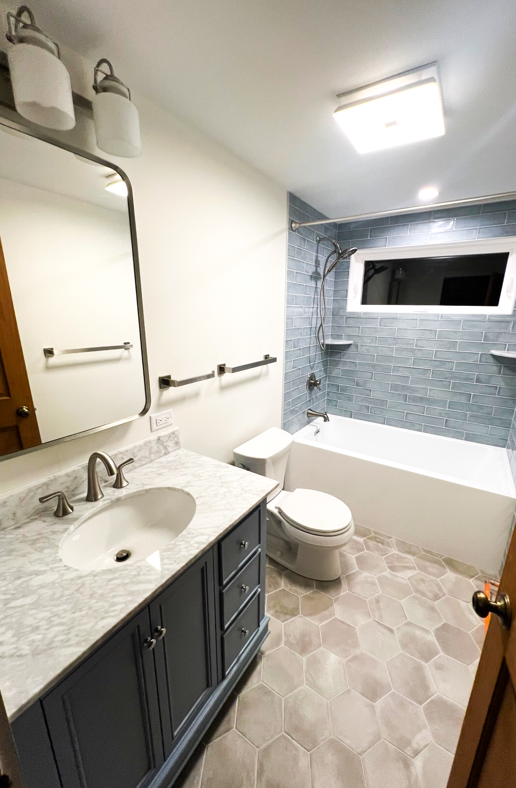 A bathroom with a marble top vanity, a mirror, a toilet, and a bathtub with a blue tile wall and a window.