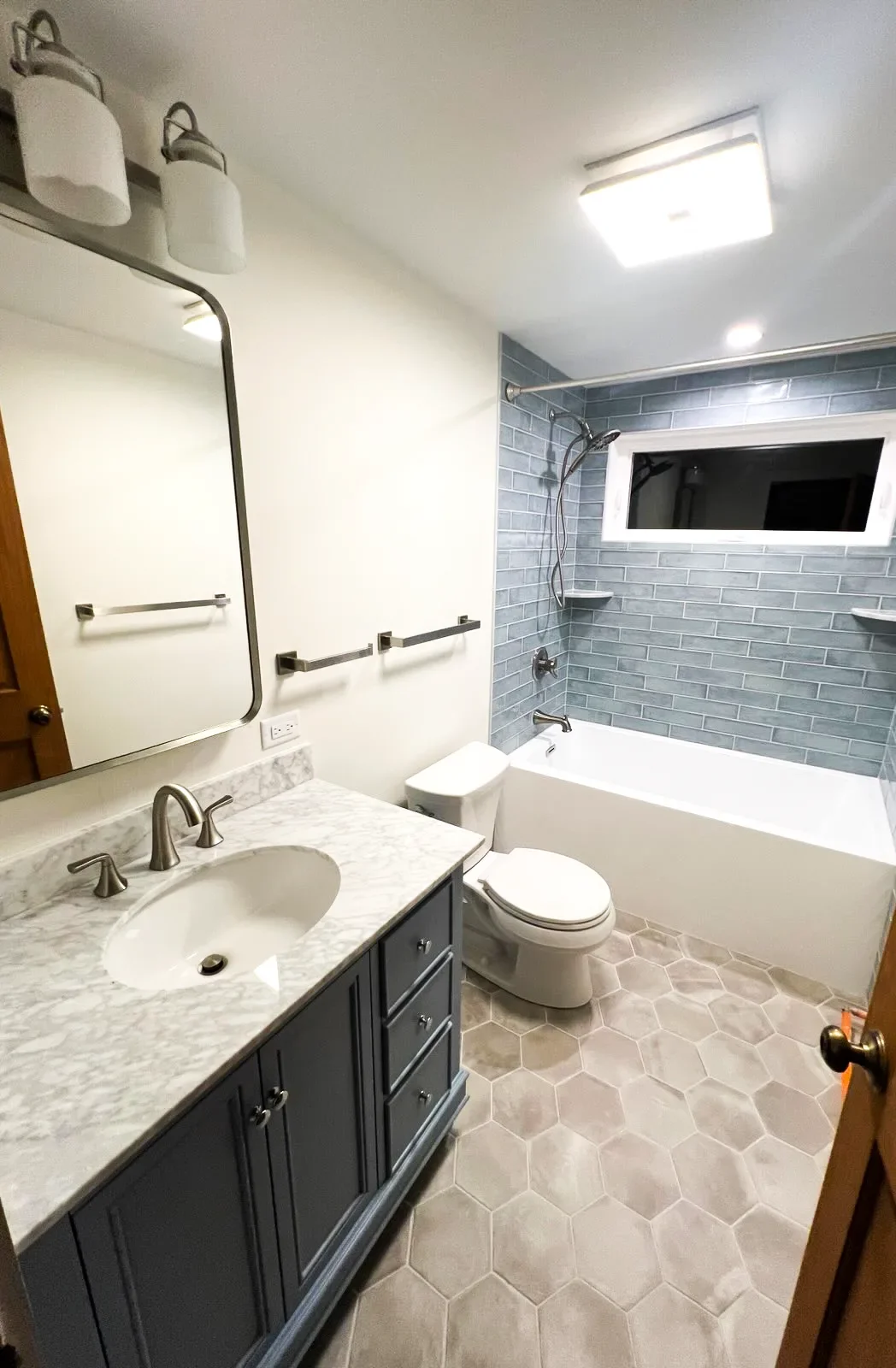 Bathroom with white walls, marble countertop vanity with dark gray cabinetry, oval sink, modern faucet, large mirror, white toilet, bathtub with gray subway tile surround, small window, hexagonal tile flooring, ceiling light, and towel racks.