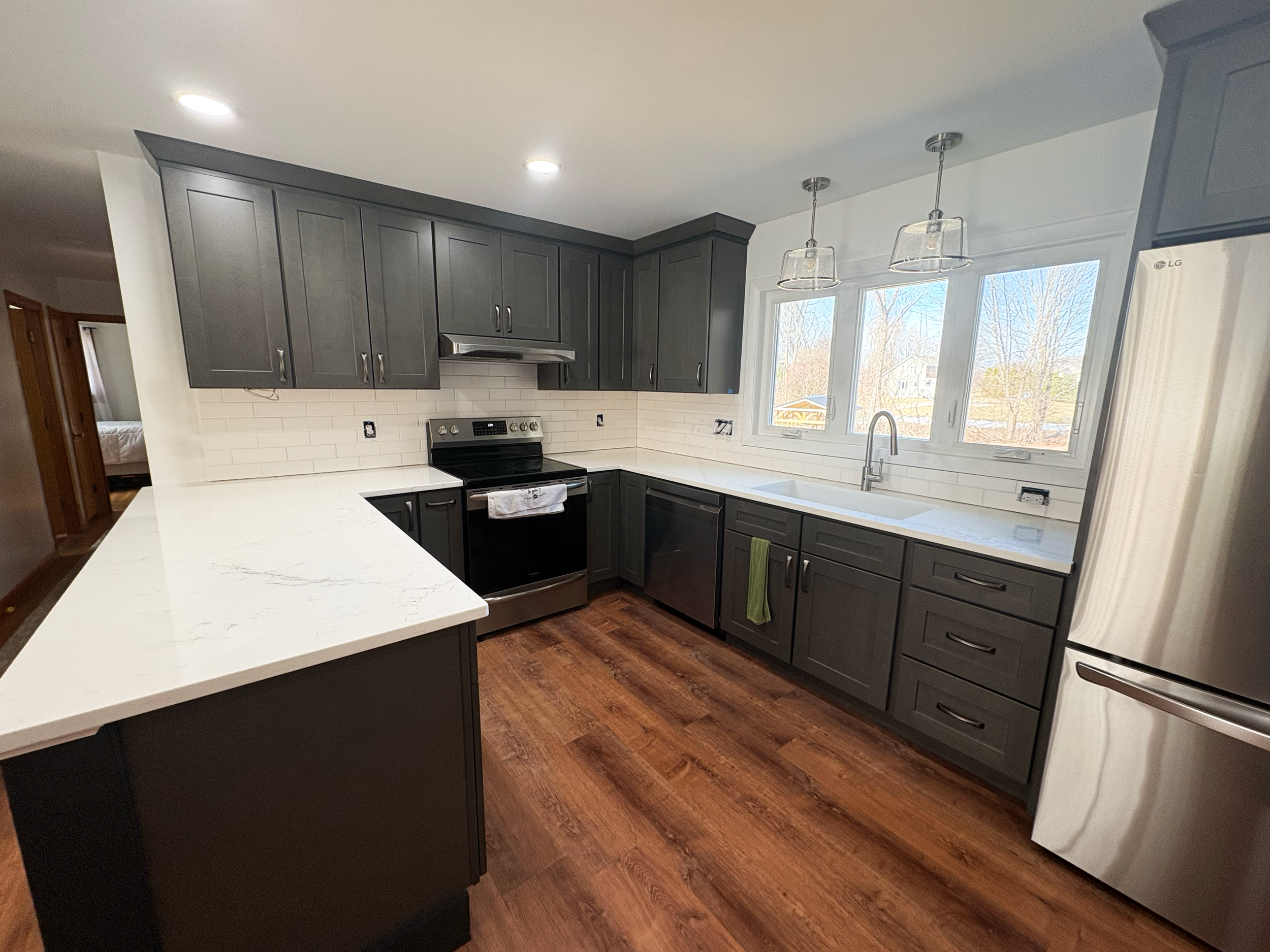 Modern kitchen with dark gray cabinets, white countertop, stainless steel appliances, large window, wooden flooring, pendant lighting, and a view of trees outside.