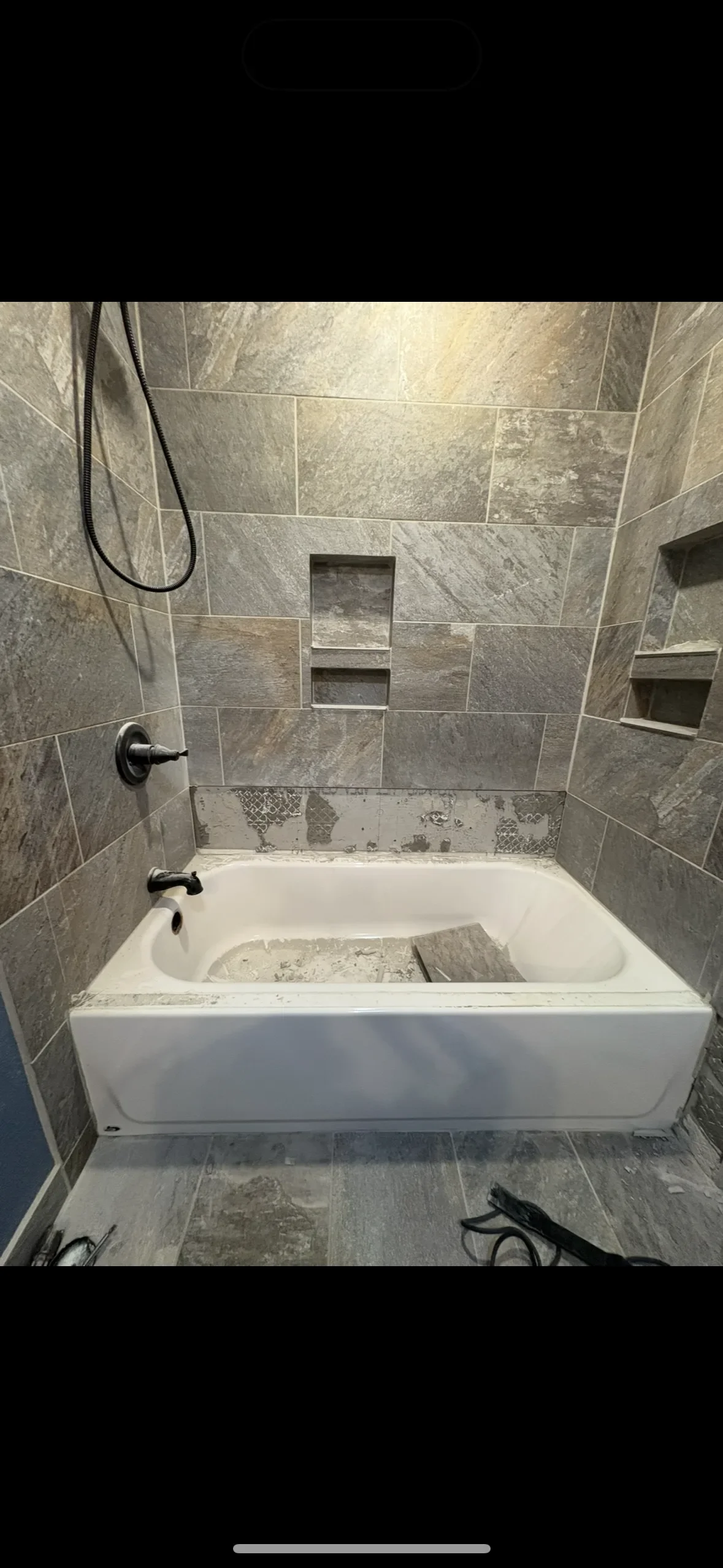 Bathroom with a bathtub under renovation, gray tiled walls with built-in shelves, missing fixtures, and construction tools on the floor.