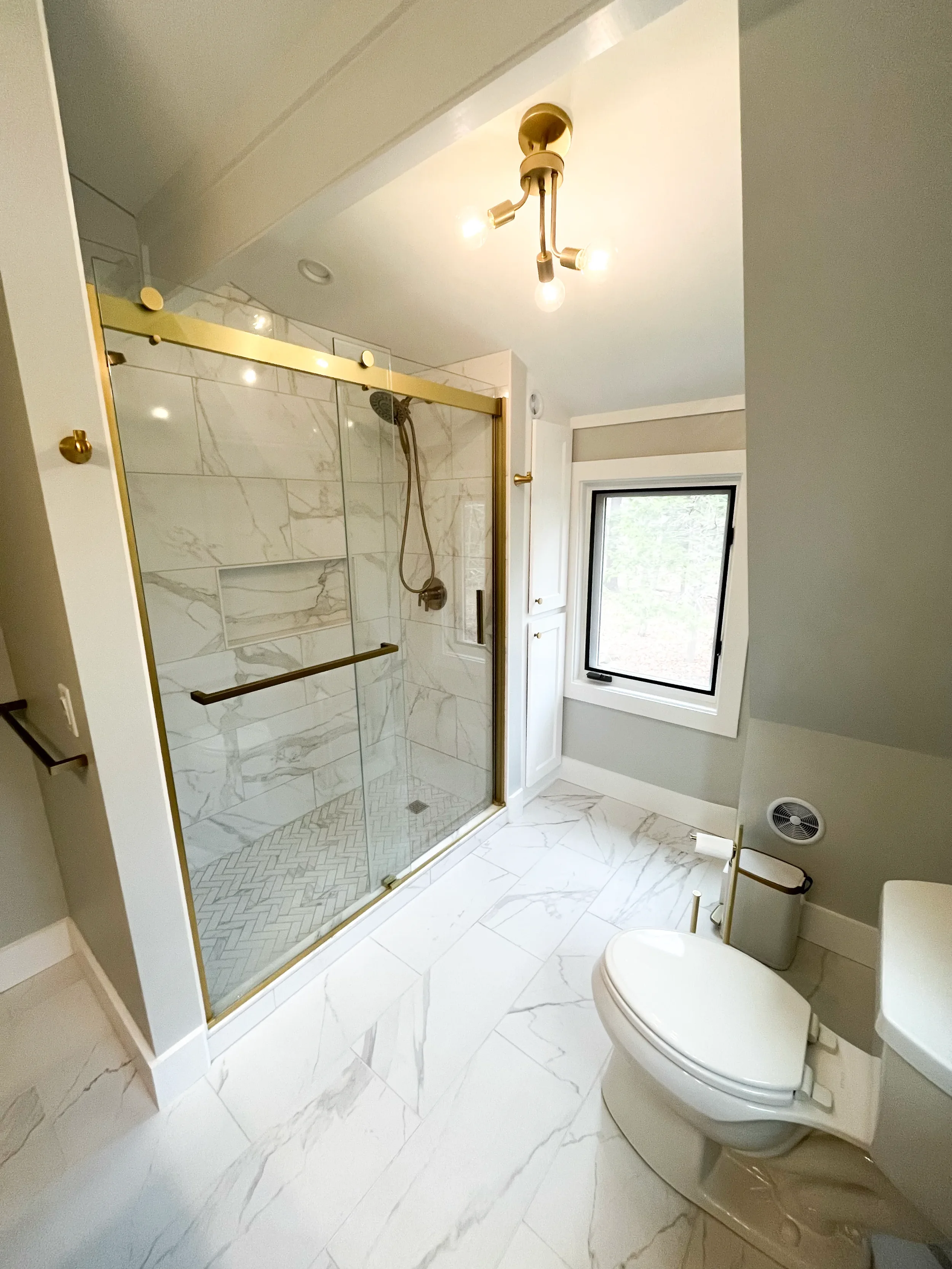 Modern bathroom with marble tile flooring, a glass shower with gold framing, a window, and a toilet.