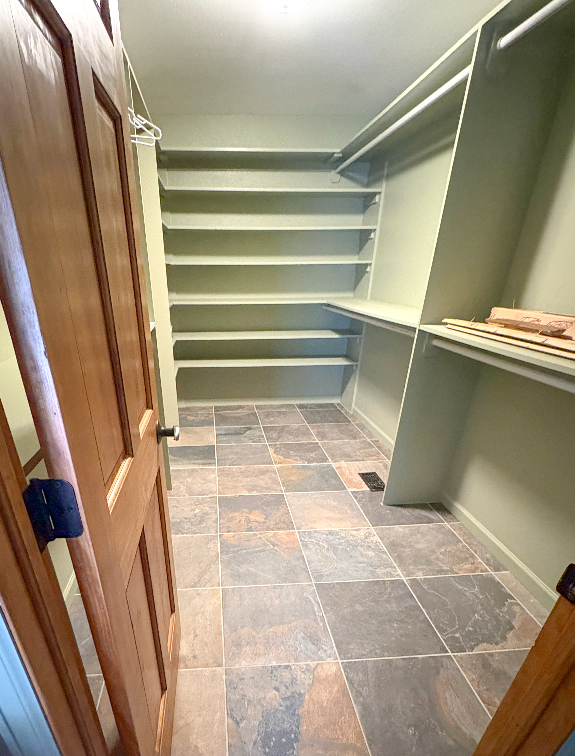 An empty walk-in closet with multiple shelves and hanging rods, featuring a wooden door with a door handle, and tiled flooring.