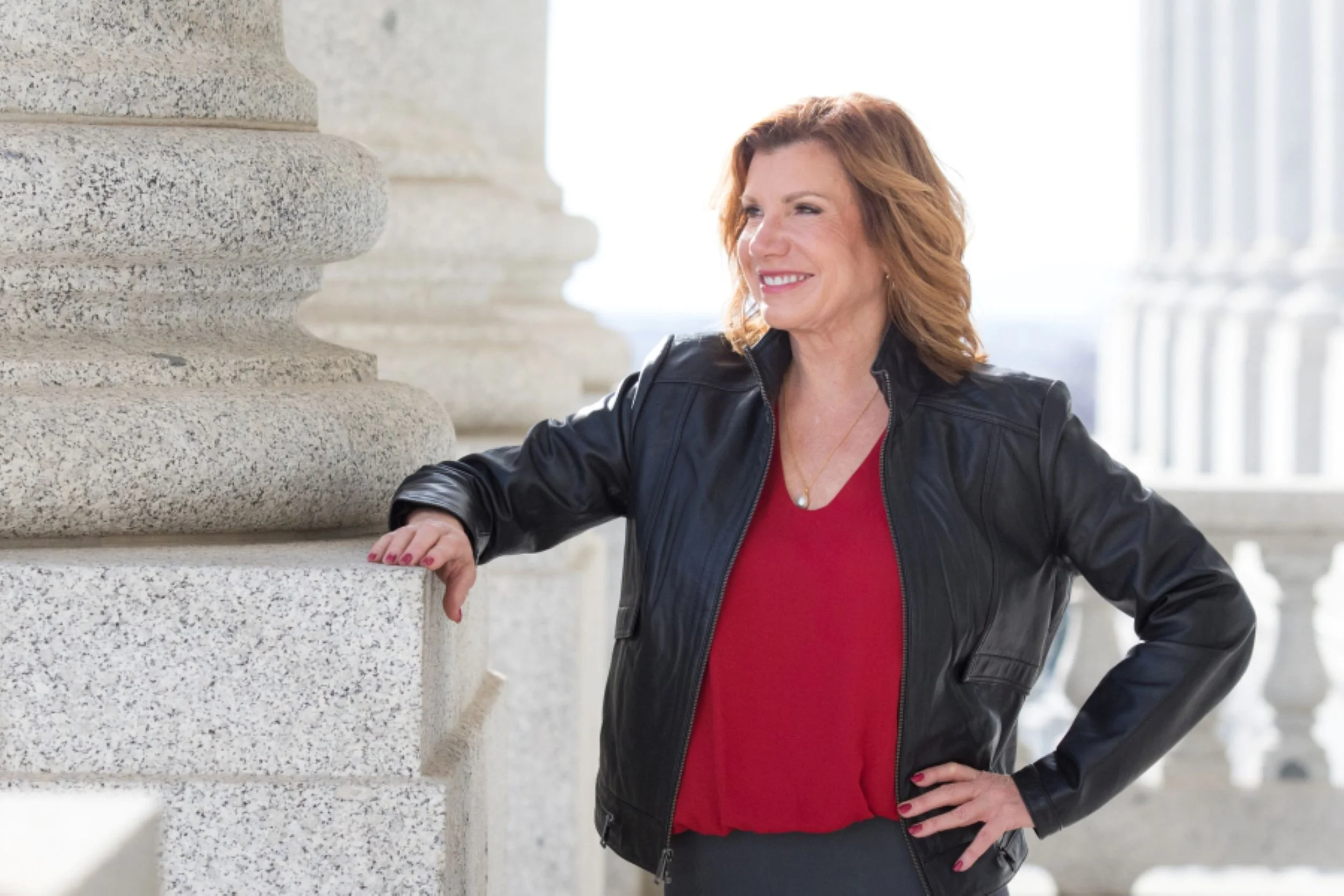 Maureen farrell in red blouse and black jacket hand on marble column smiling while looking into distance