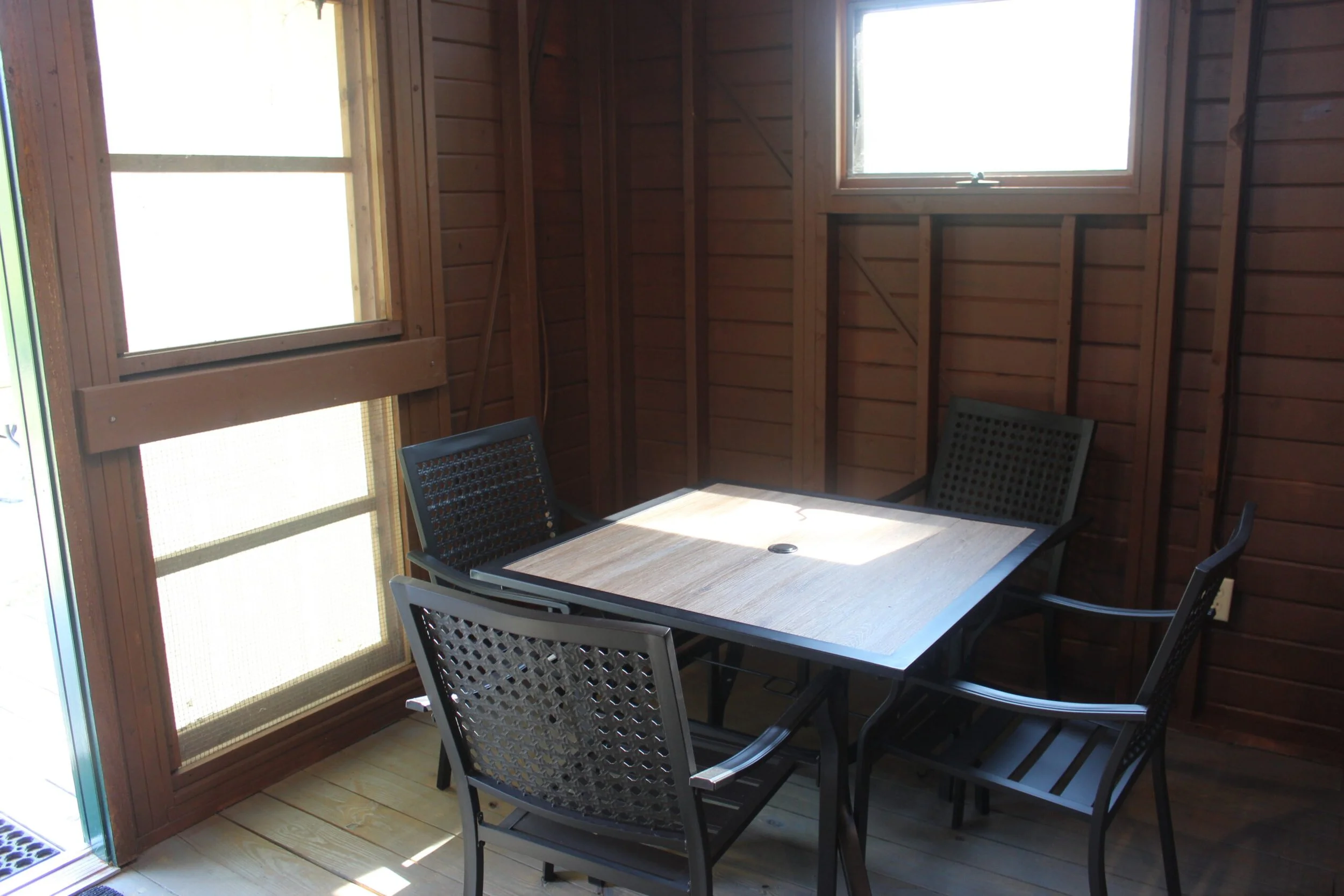 A small outdoor screened-in patio with wooden walls and ceiling, featuring a rectangular table with four black chairs around it. There are two windows in the background and a sliding glass door on the left side.