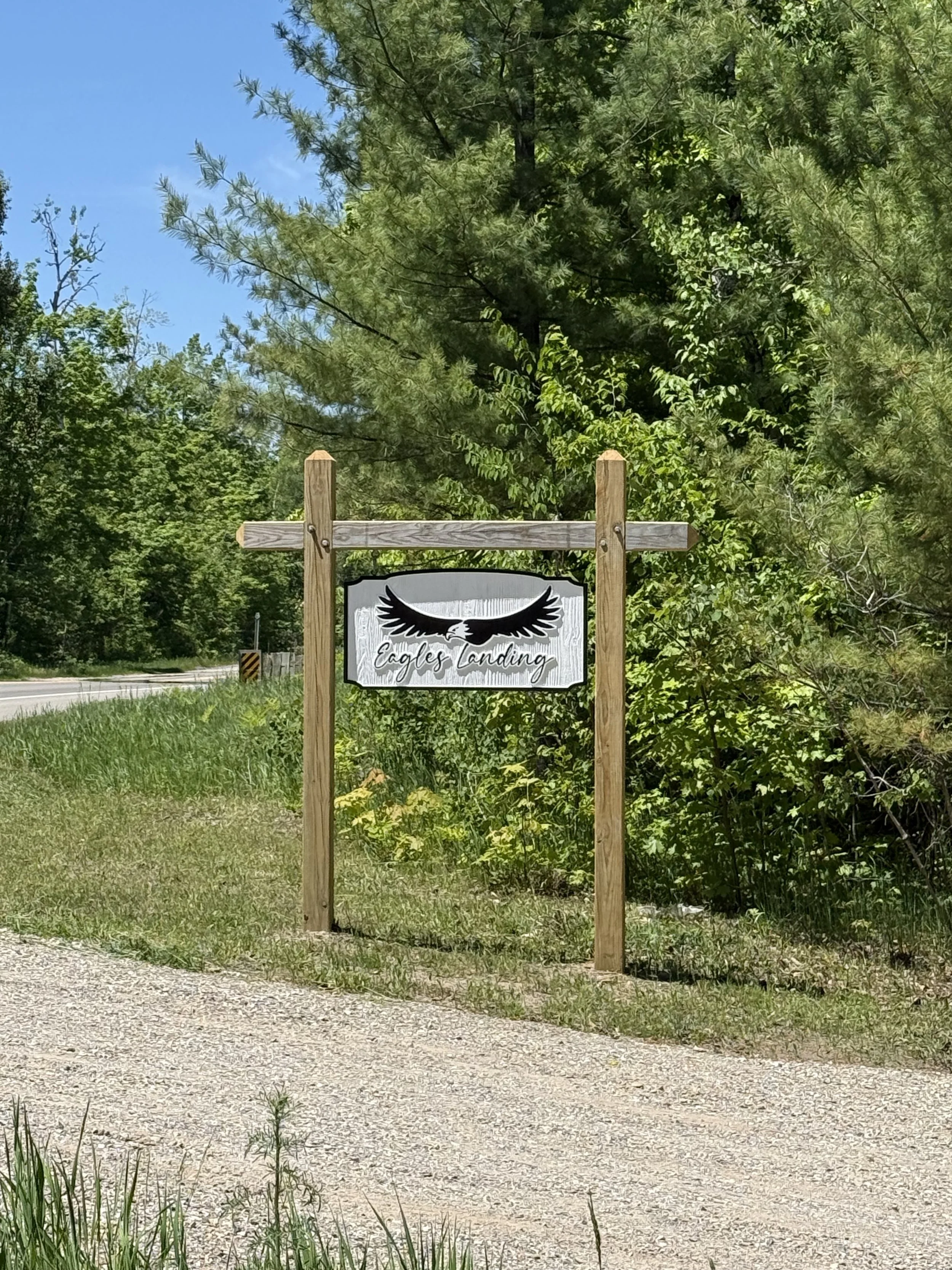 A sign with the logo of black wings and the text 'Eagles Landing' on it, standing on wooden posts beside a tree with green leaves, in a natural outdoor setting.