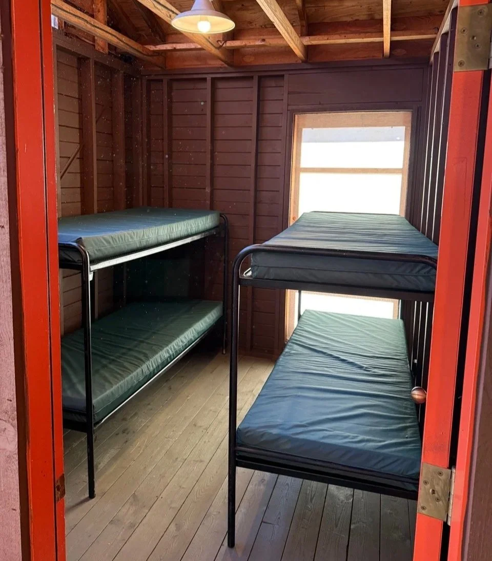 Inside a small wooden room with four metal bunk beds with green mattresses, two on each side, and a window at the back, illuminated by a ceiling light.